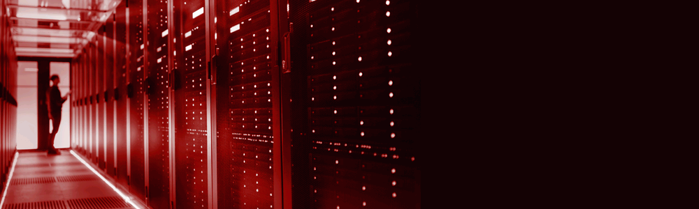 A person standing in a hallway lined with red server racks and blinking lights, conveying a data center or technology environment.