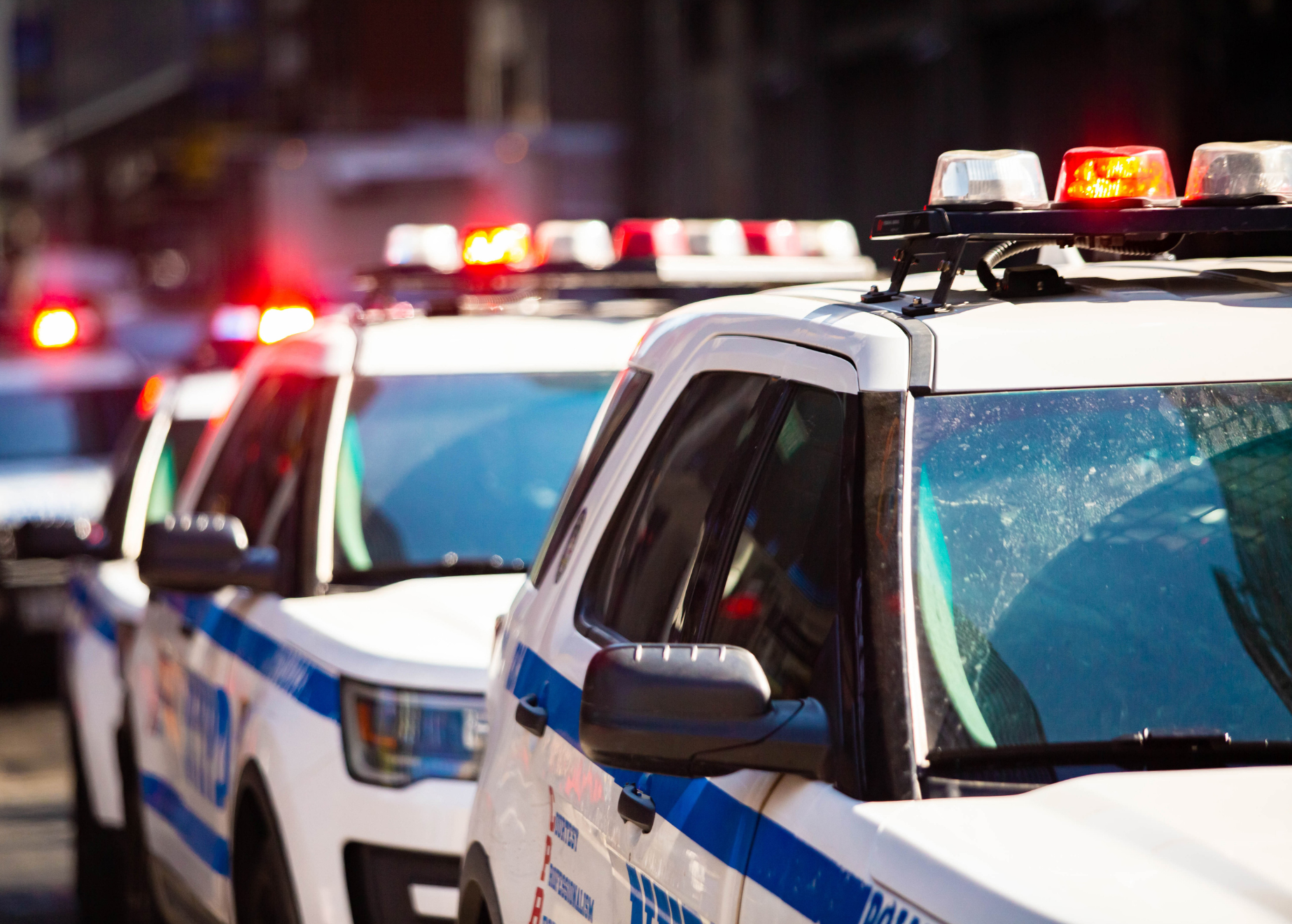 Line of police vehicles with flashing red and white emergency lights parked outdoors.