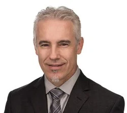 Professional headshot of Co-Founder, Clifford Dice wearing a dark suit, patterned tie, and light shirt, against a plain white background.