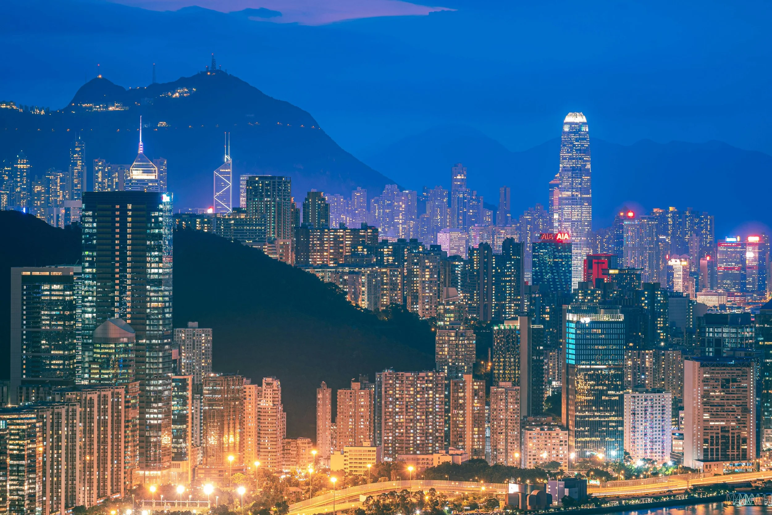 Night view of a city skyline with tall skyscrapers, illuminated office buildings, and a mountain in the background, under a dusk sky.