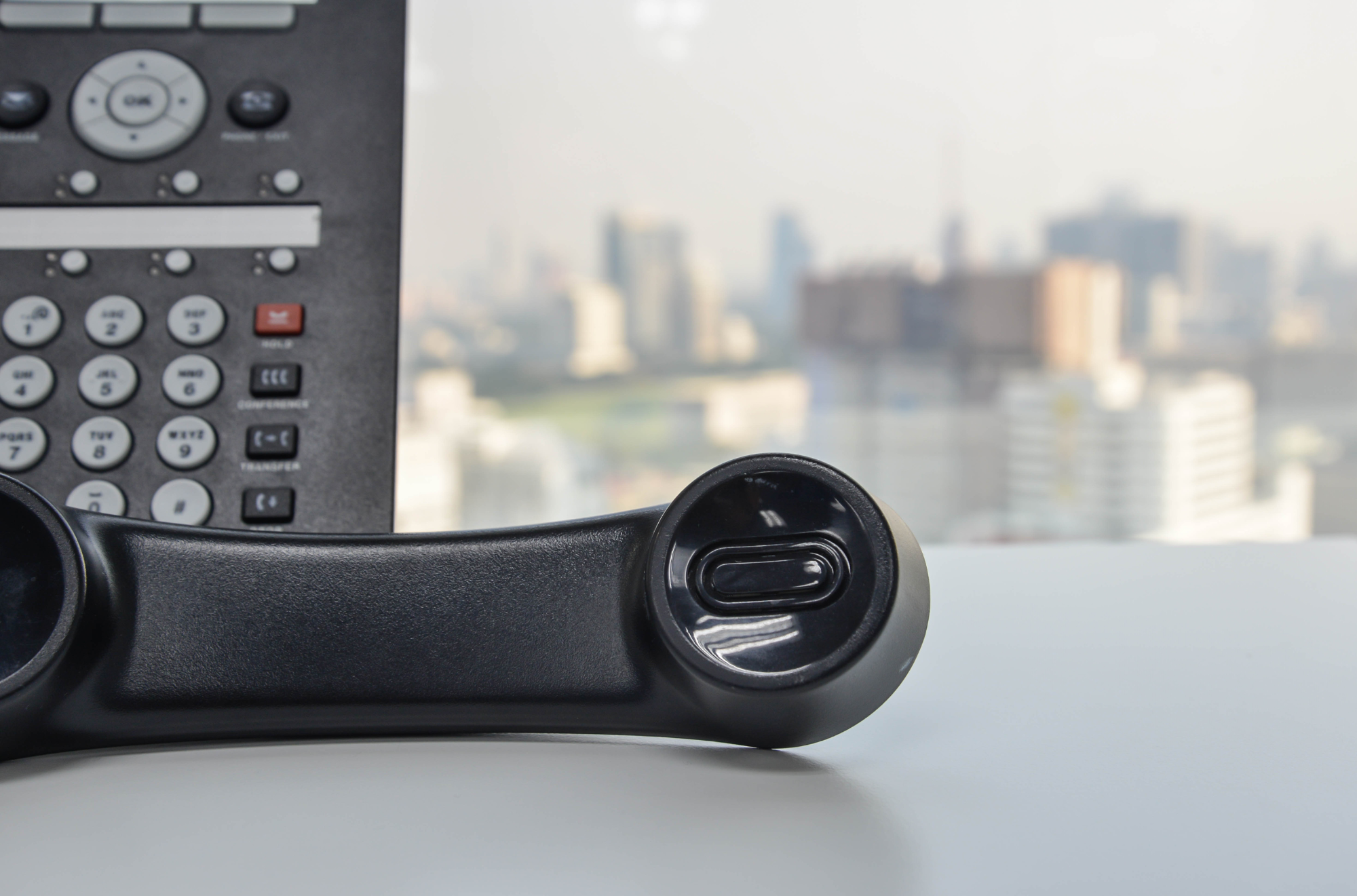 Close-up of a telephone handset on a white desk with a blurred cityscape background visible through a window.