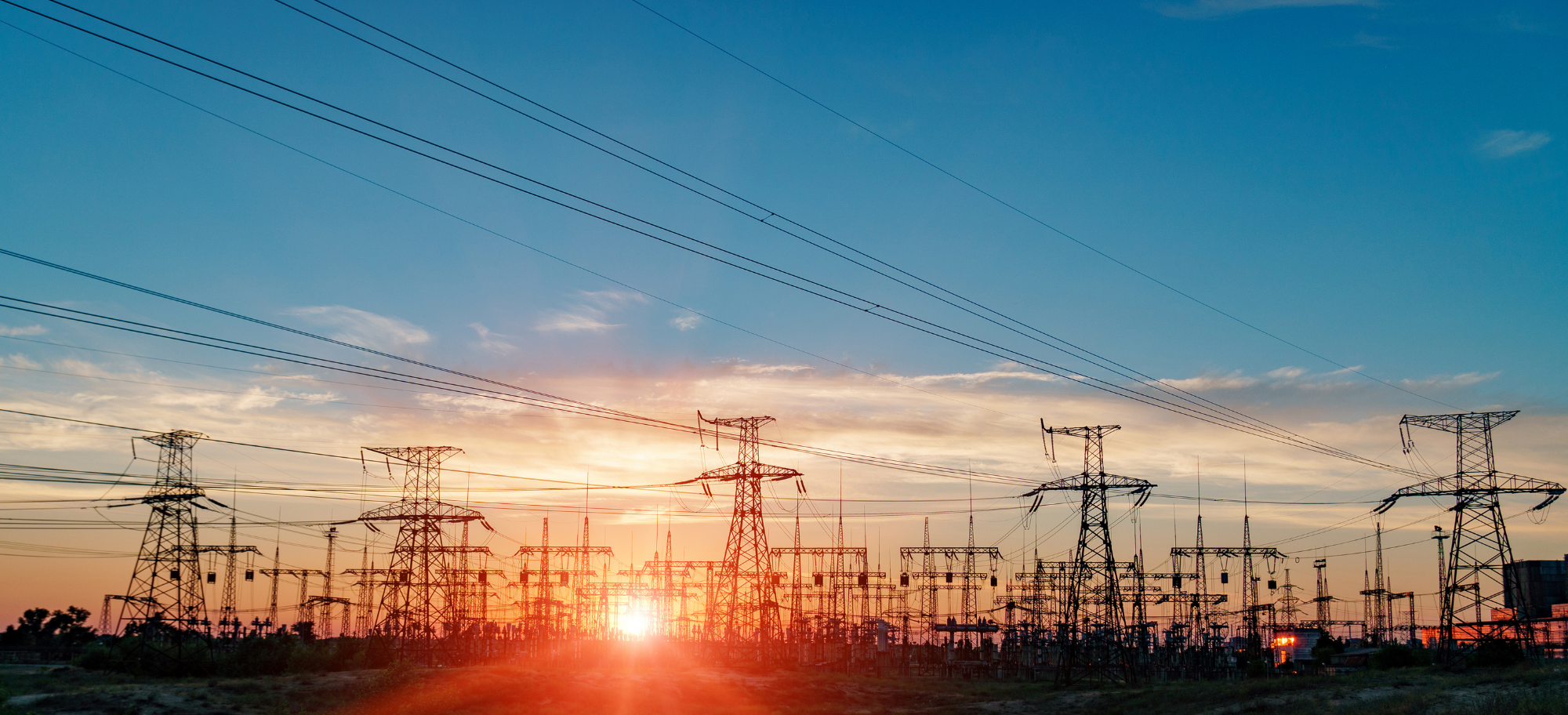 Sunset over a large power plant with multiple electrical transmission towers and power lines against the sky.