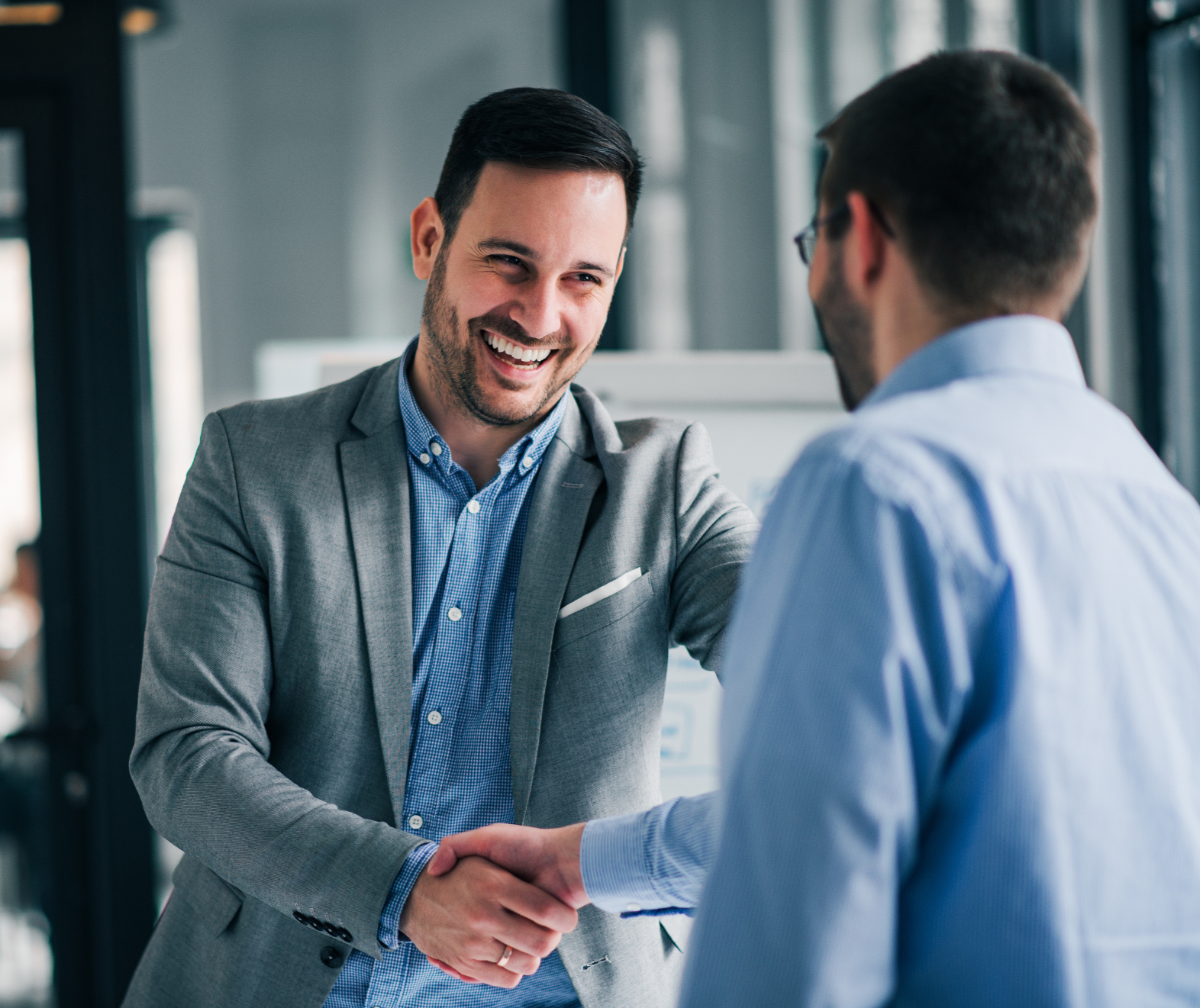 Two men shaking hands in a business setting, one is wearing a gray blazer and blue checkered shirt, smiling, the other is wearing glasses and a light blue shirt.