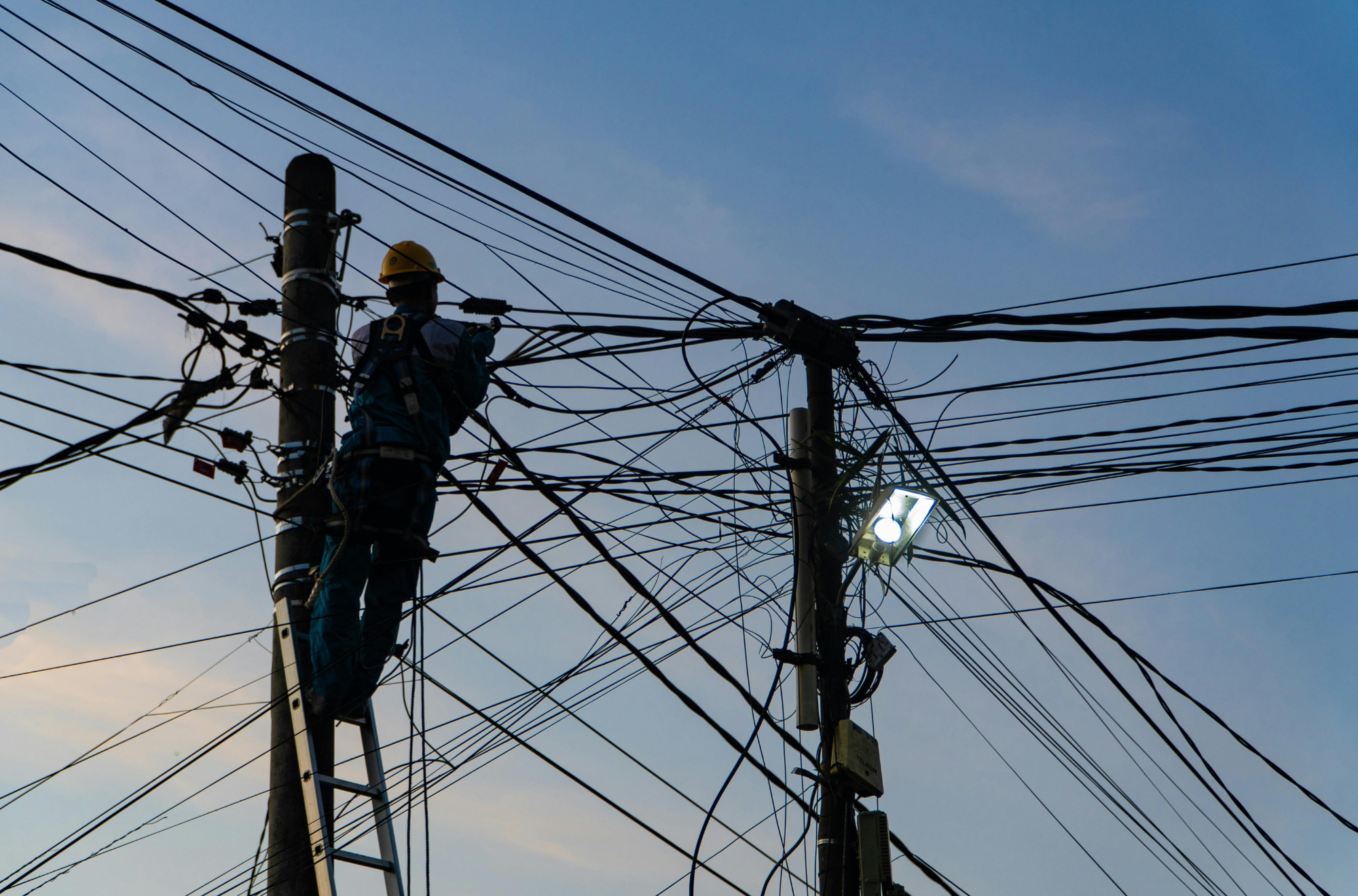 A worker wearing a yellow safety helmet working on electric power lines on a utility pole at dusk or early evening with a clear sky in the background.