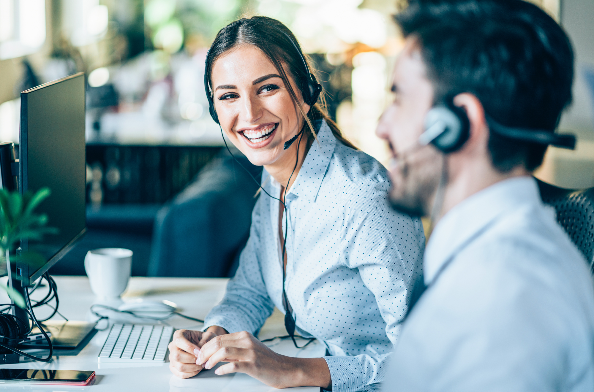 Two call center agents, a woman and a man, are sitting at desks with computer monitors and wearing headsets, engaged in a conversation in an office environment.