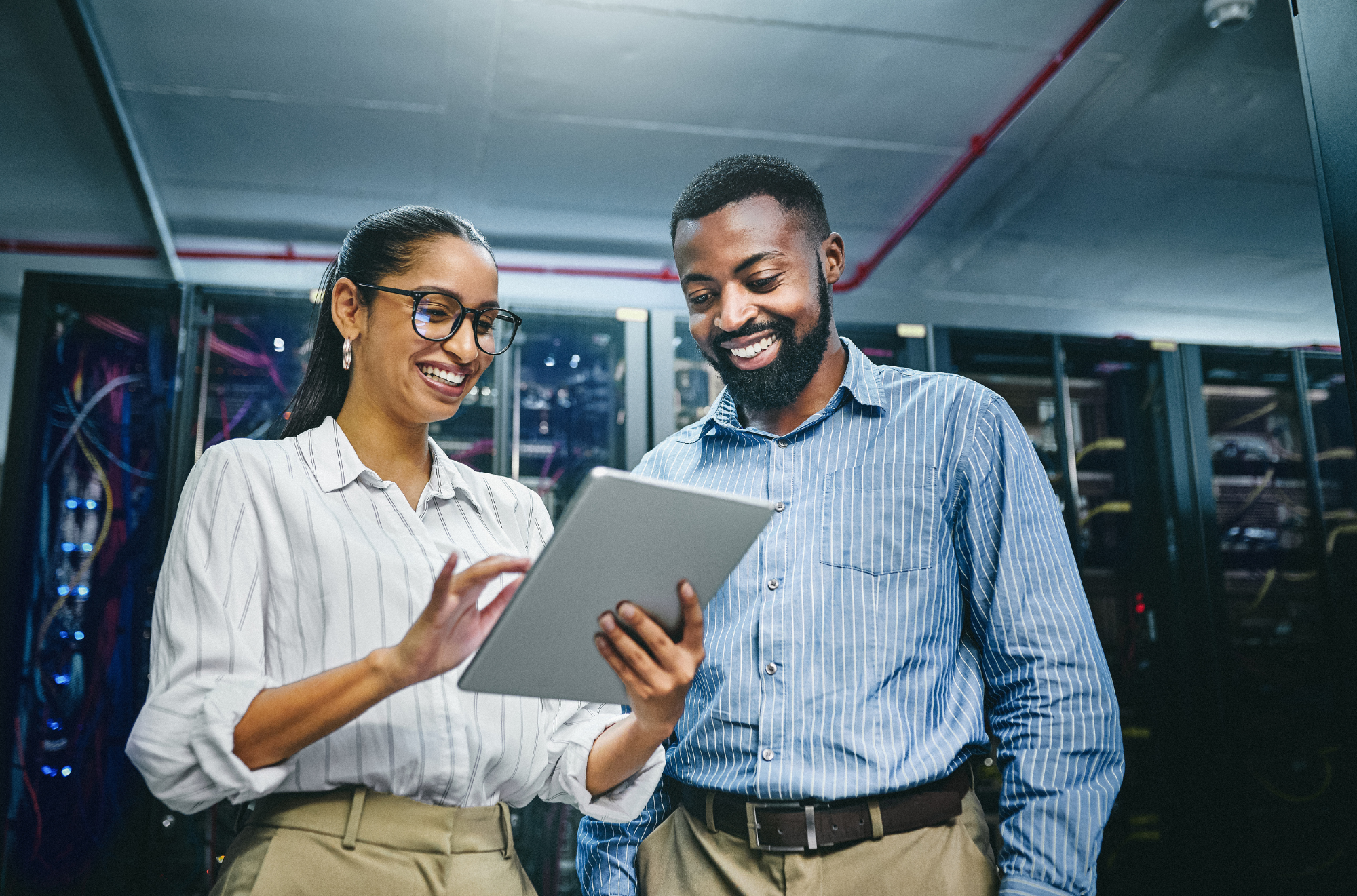 Two IT professionals, a woman and a man, standing in a server room, looking at a tablet and smiling.