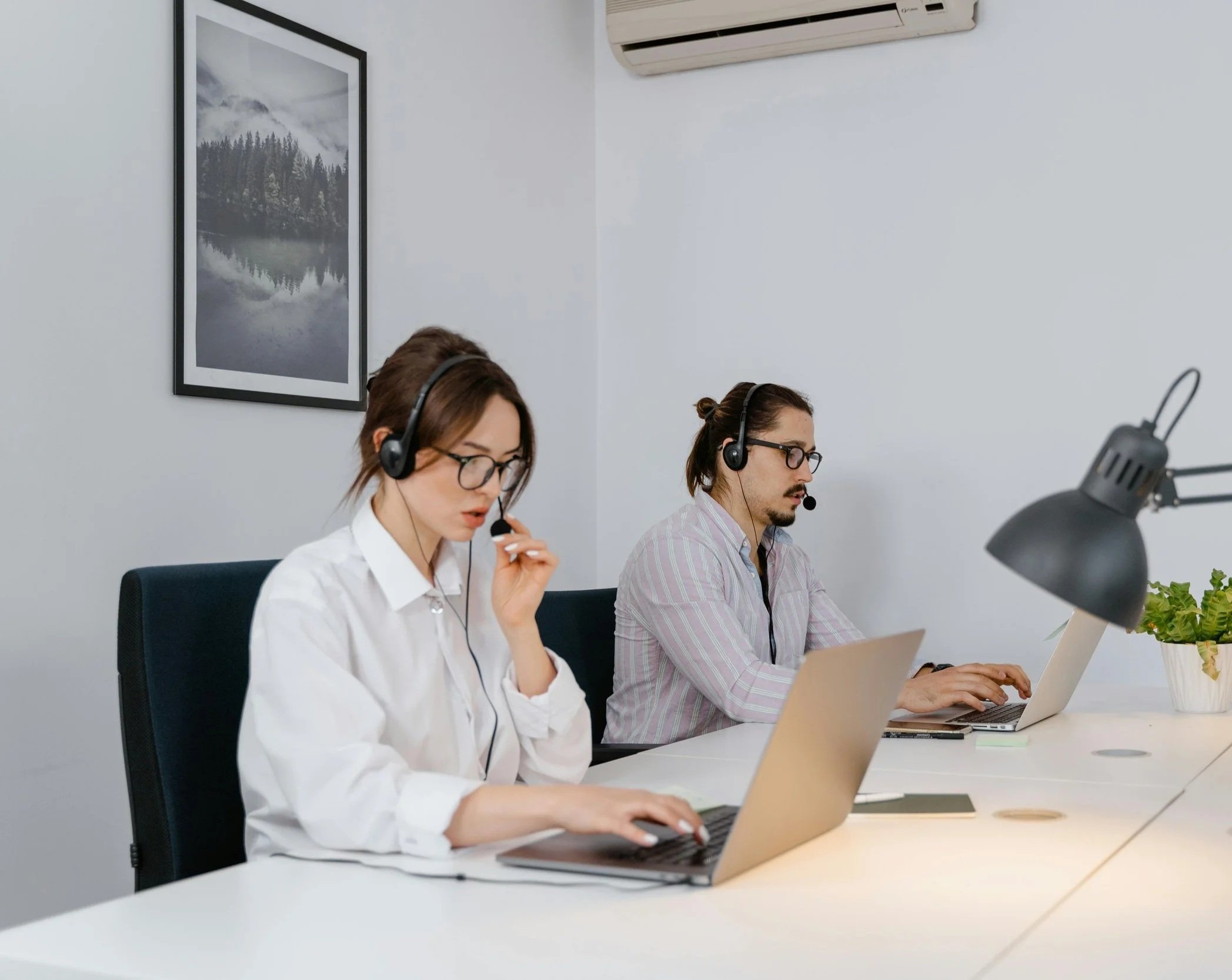 Two people in a modern office setting working at a desk, wearing headsets and using laptops, with a framed landscape photograph of mountains and a lake on the wall behind them and an air conditioning unit above.