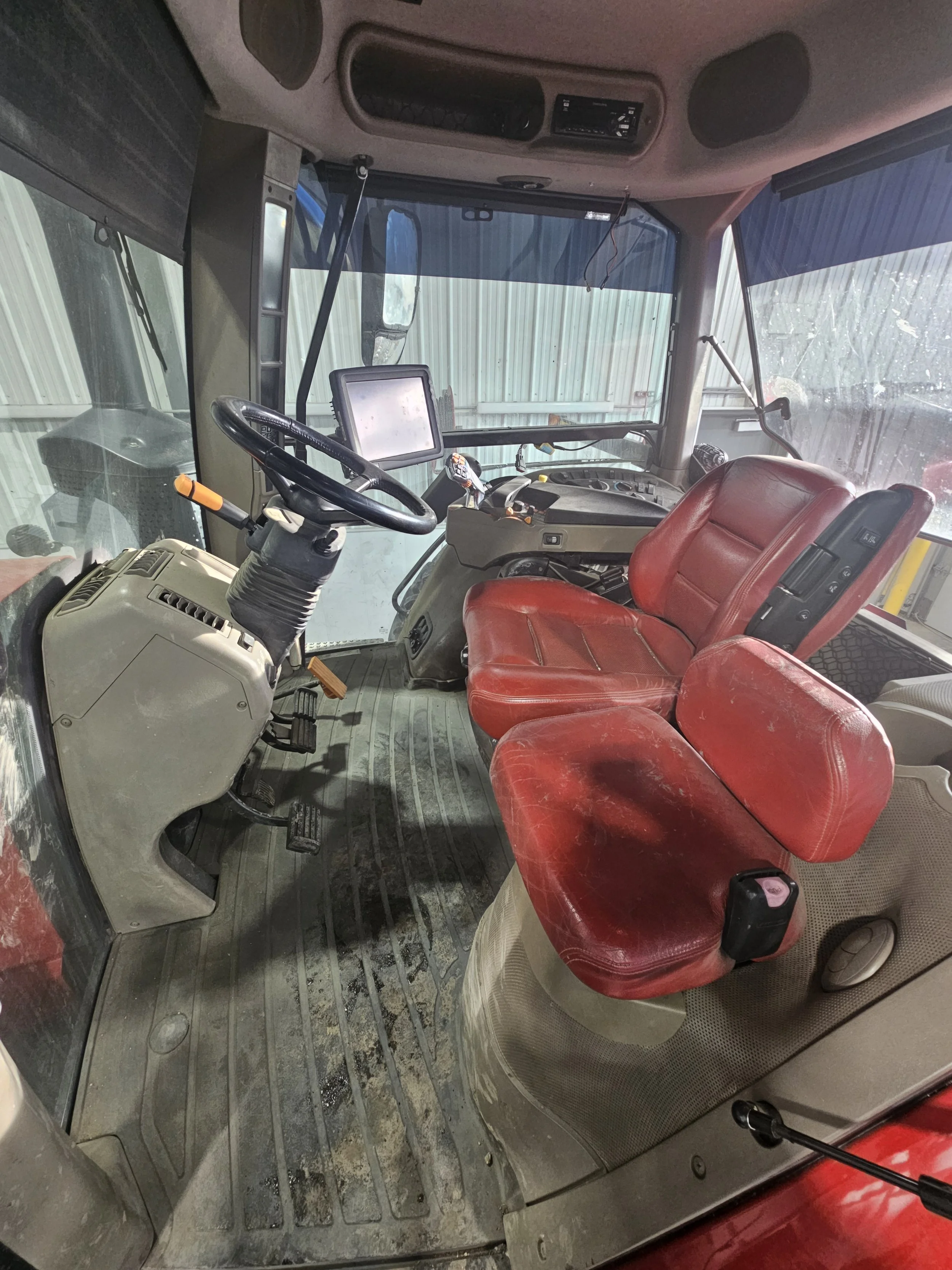 Interior of a tractor cab with red seat, steering wheel, dashboard, and control panel, with a large touchscreen display and various controls.