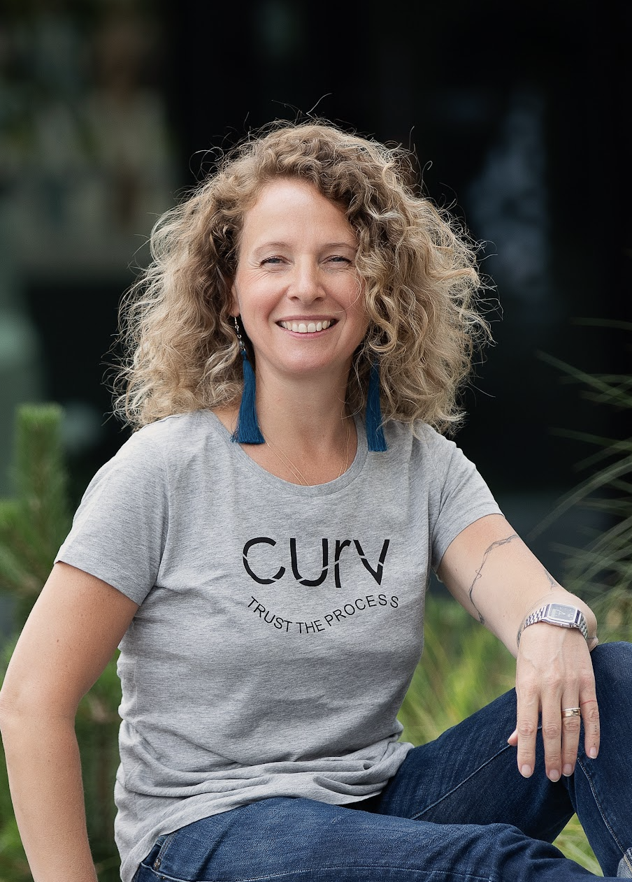 Woman with curly blonde hair wearing a gray t-shirt with 'CURV' and 'Trust the Process' written on it, sitting outdoors with greenery around.