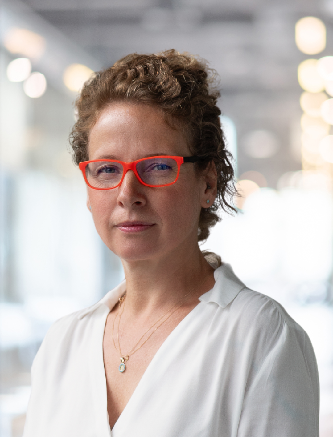 A woman with short curly hair and red glasses, wearing a white blouse, stands in a well-lit, blurred indoor environment.
