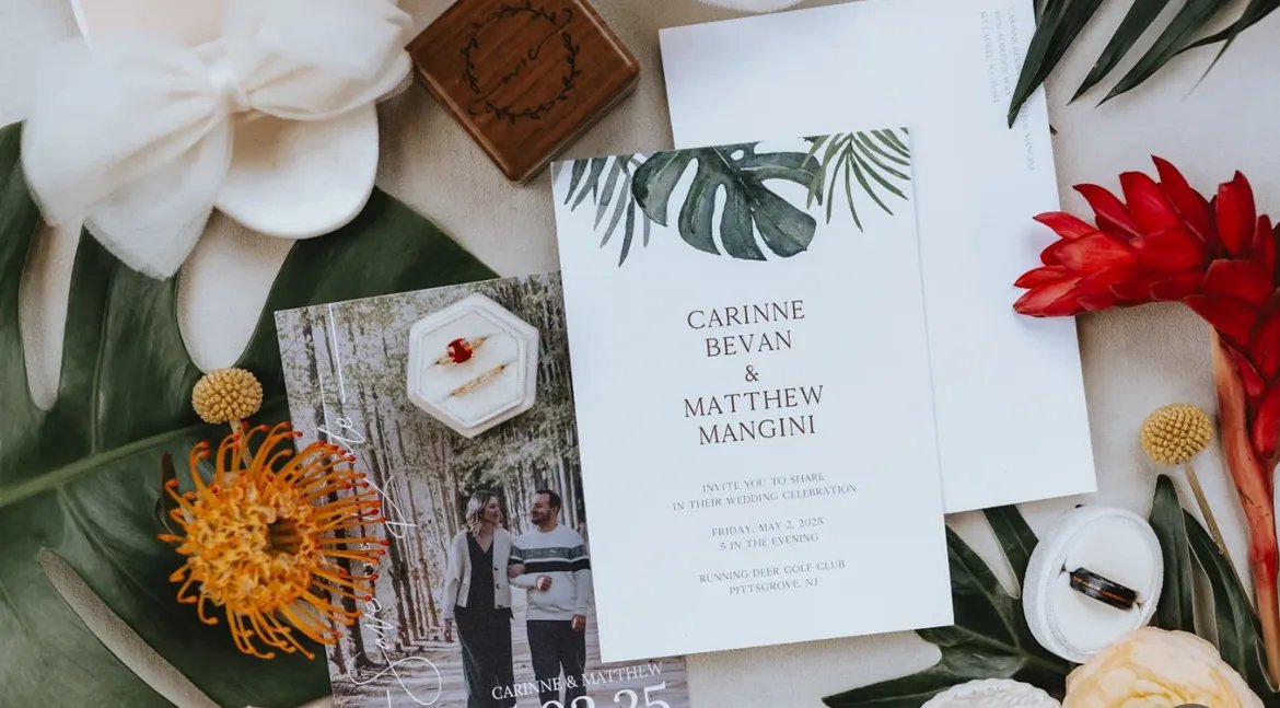 Wedding invitation with greenery design, surrounded by floral elements, rings, a photo of the couple, and jewelry on a table.