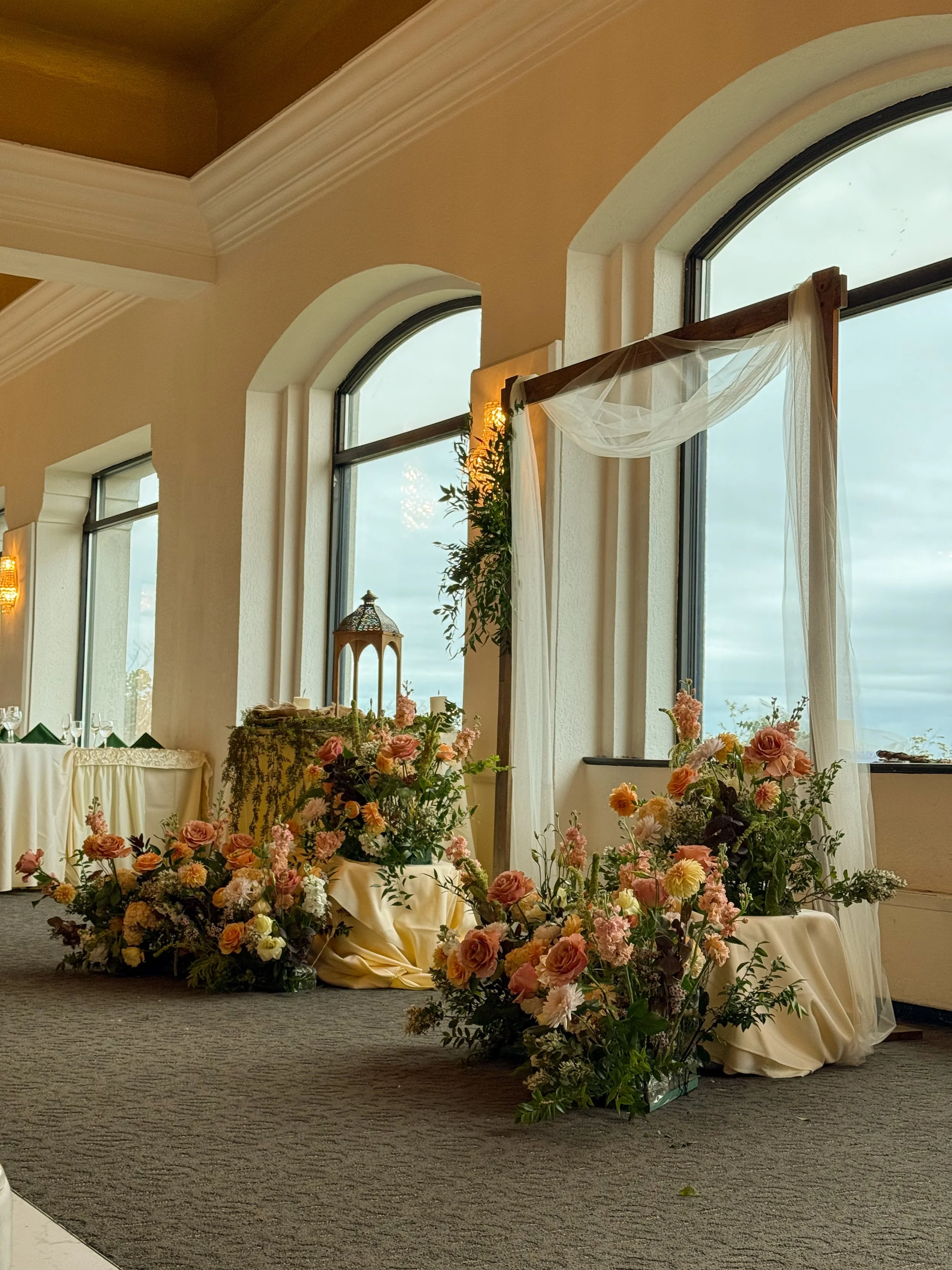 Elegant wedding or event decorations with large floral arrangements and arch in soft pink, peach, and cream colors, set on cream-colored tablecloths near tall, arched windows with sheer drapes in a spacious, well-lit room.