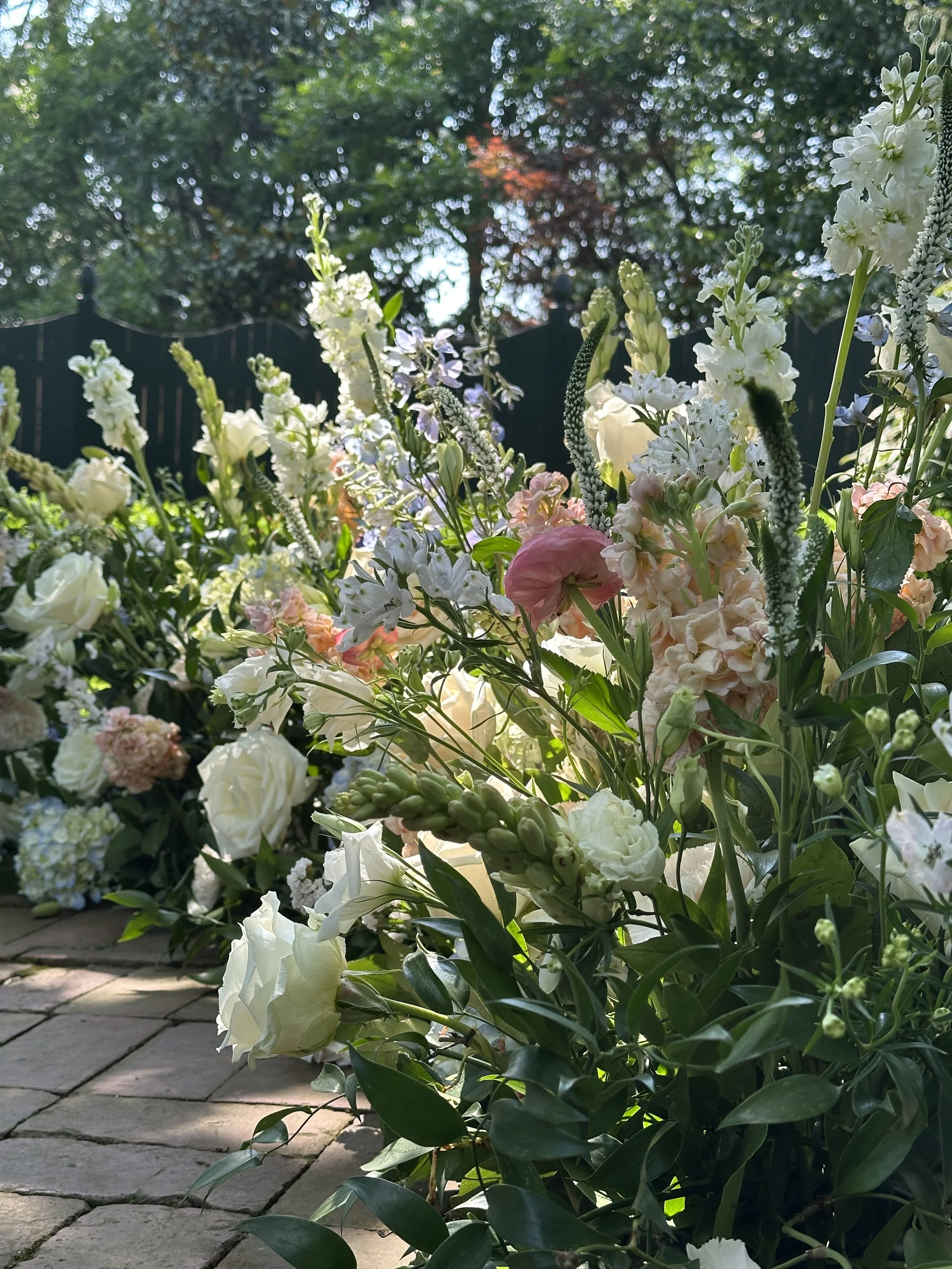 A close-up view of garden style flower arrangement with white and pale pink flowers, with a black fence and trees in the background; sunlight filters through the trees.