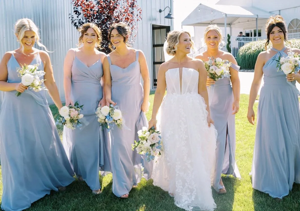 Bride and five bridesmaids walking outdoors, holding bouquets, in a garden setting with a white building and tent in the background in Pitman New Jersey.