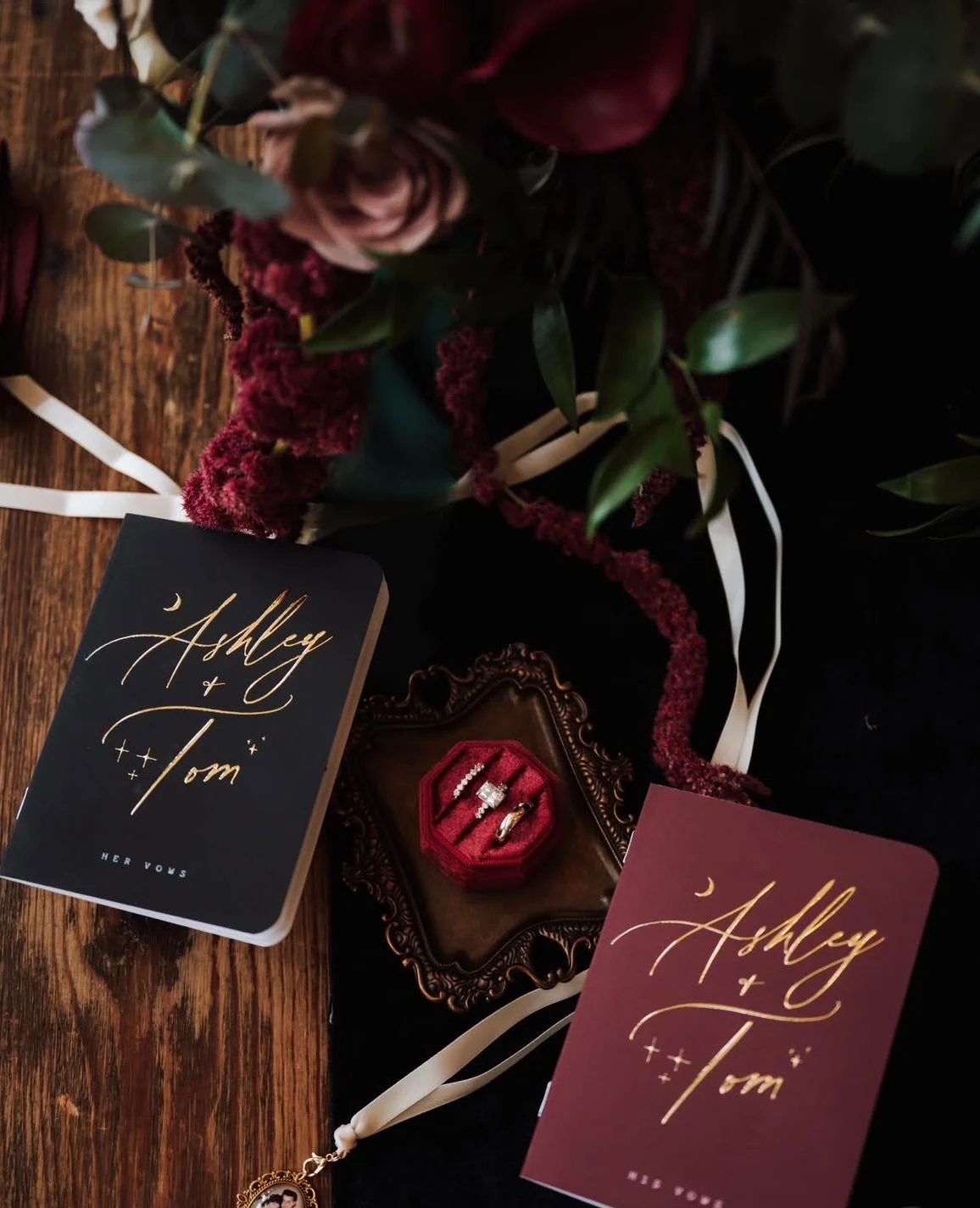 A wedding invitation display on a moody surface with flowers in the background, a jewelry box with rings, wedding vows with gold lettering, and a wedding ring Running Deer New Jersey.