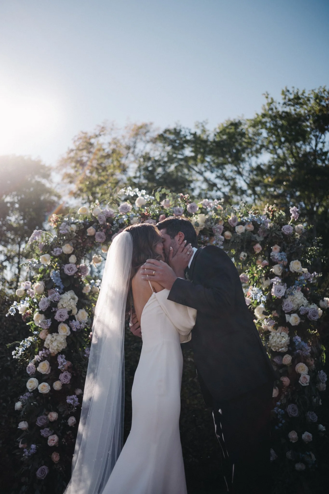 A couple kissing at their wedding under a floral arch with sunlight shining through trees in the background in Cape May New Jersey.
