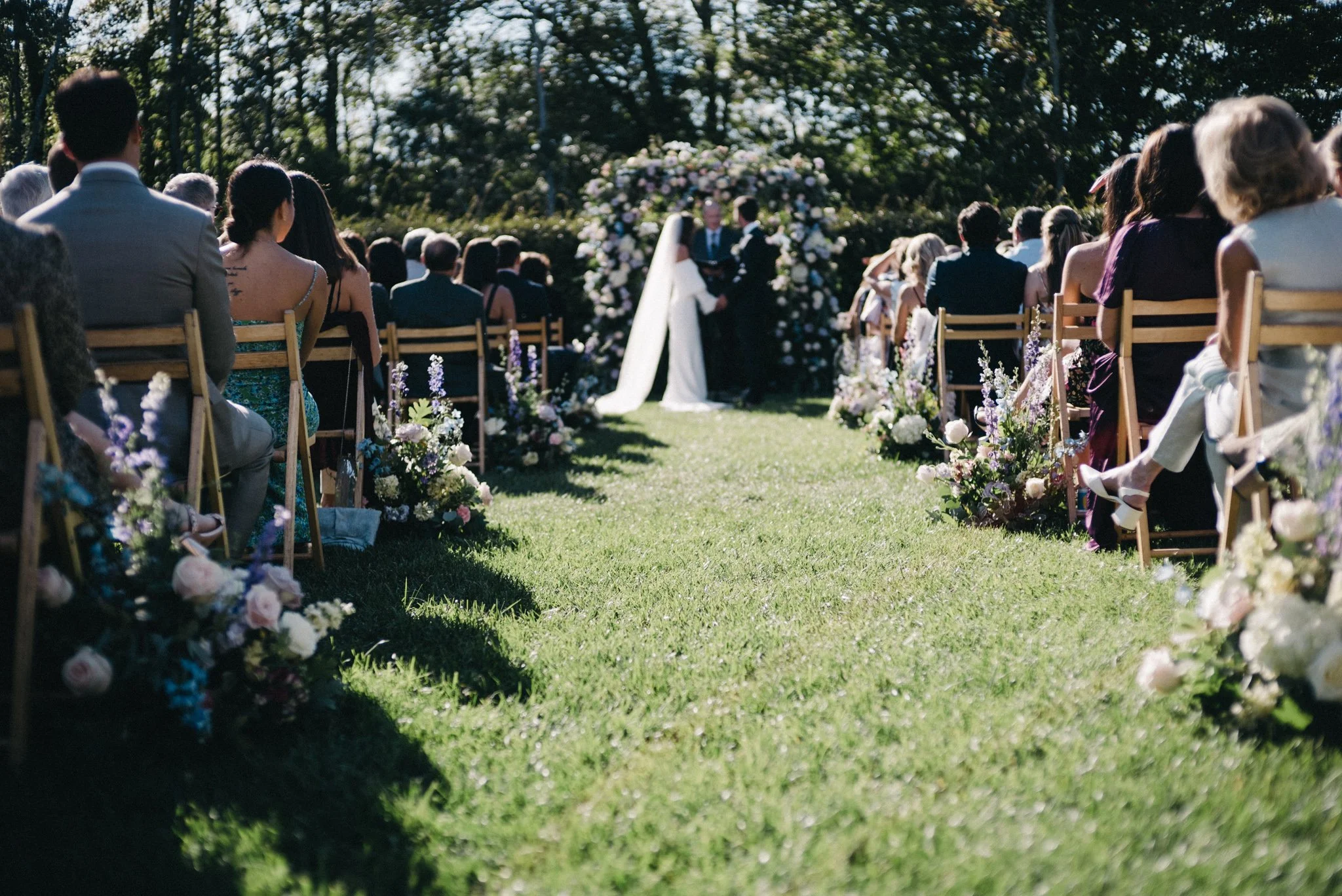 Outdoor wedding ceremony with the bride and groom standing under a floral arch, surrounded by guests seated on wooden chairs decorated with flowers in Cape May New Jersey.