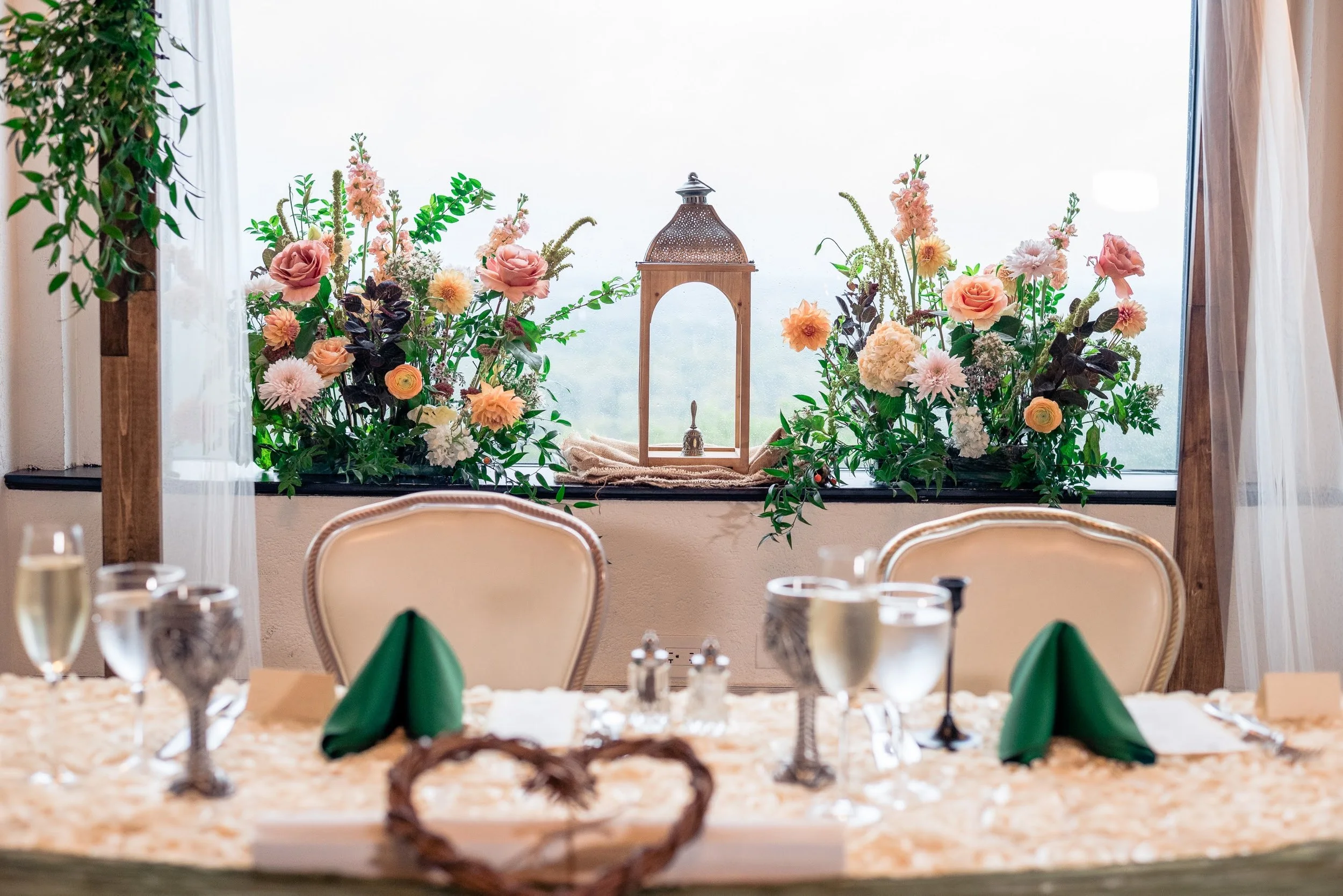 A decorated sweetheart table with green napkins, wine glasses, and silverware, with a floral centerpiece and window with pink and peach-colored flowers and greenery in the background overlooking New York City in North Jersey.