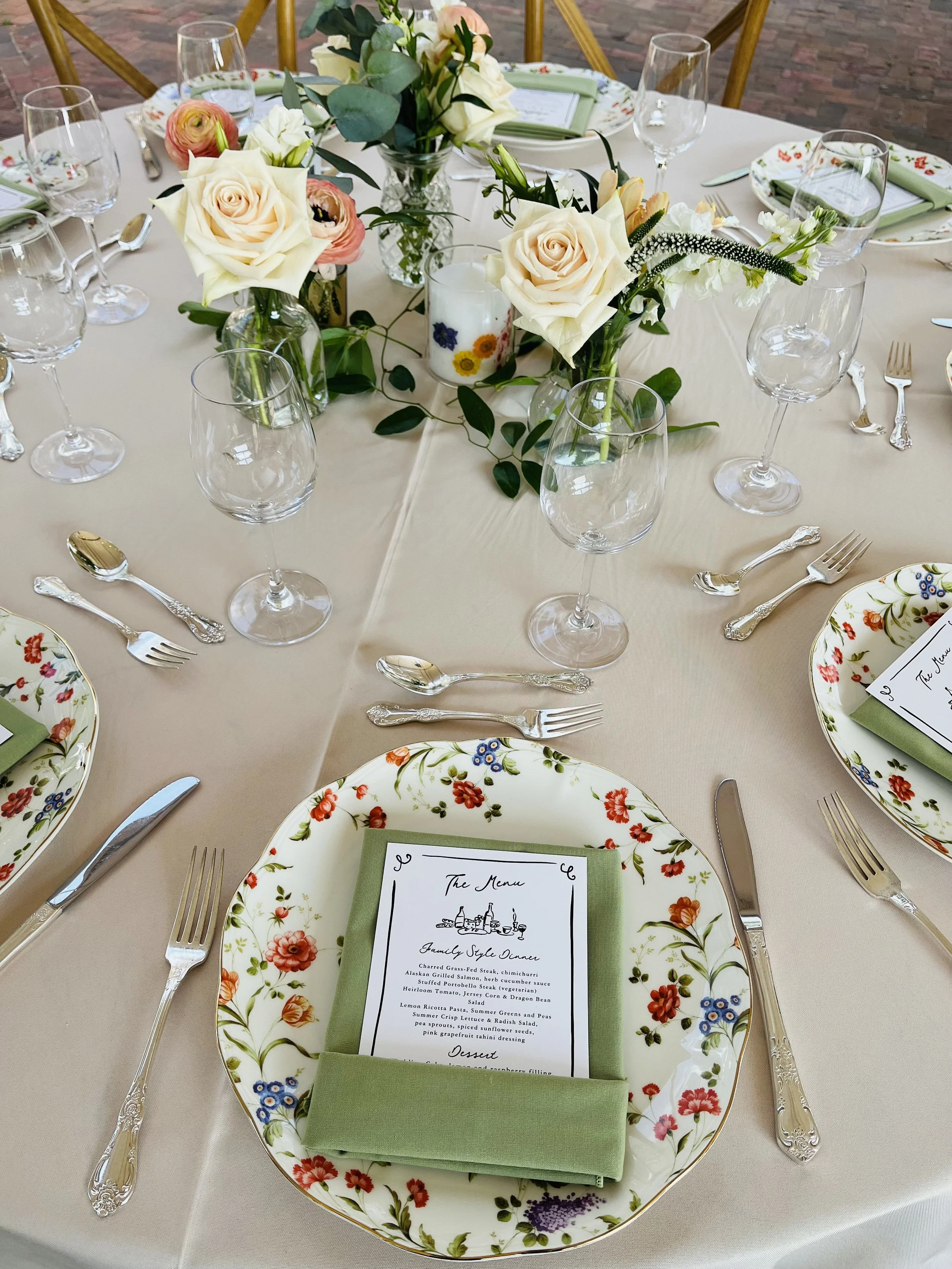 A round dining table set with floral-patterned plates, silverware, wine glasses, green napkins, and a floral centerpiece with white and pink roses and greenery.