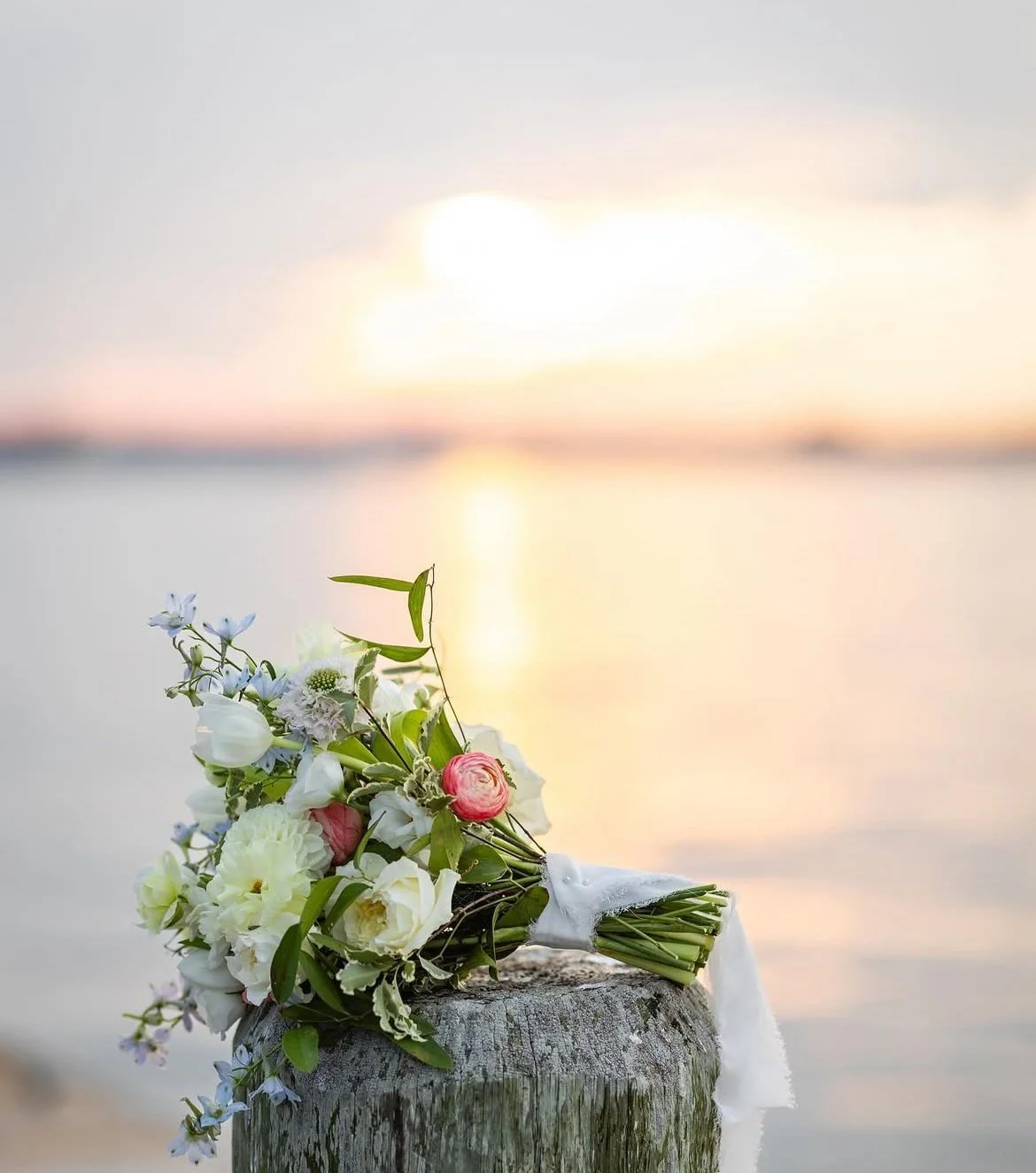 A bouquet of white, pink, and purple flowers resting on a weathered wooden post at the beach during sunset.