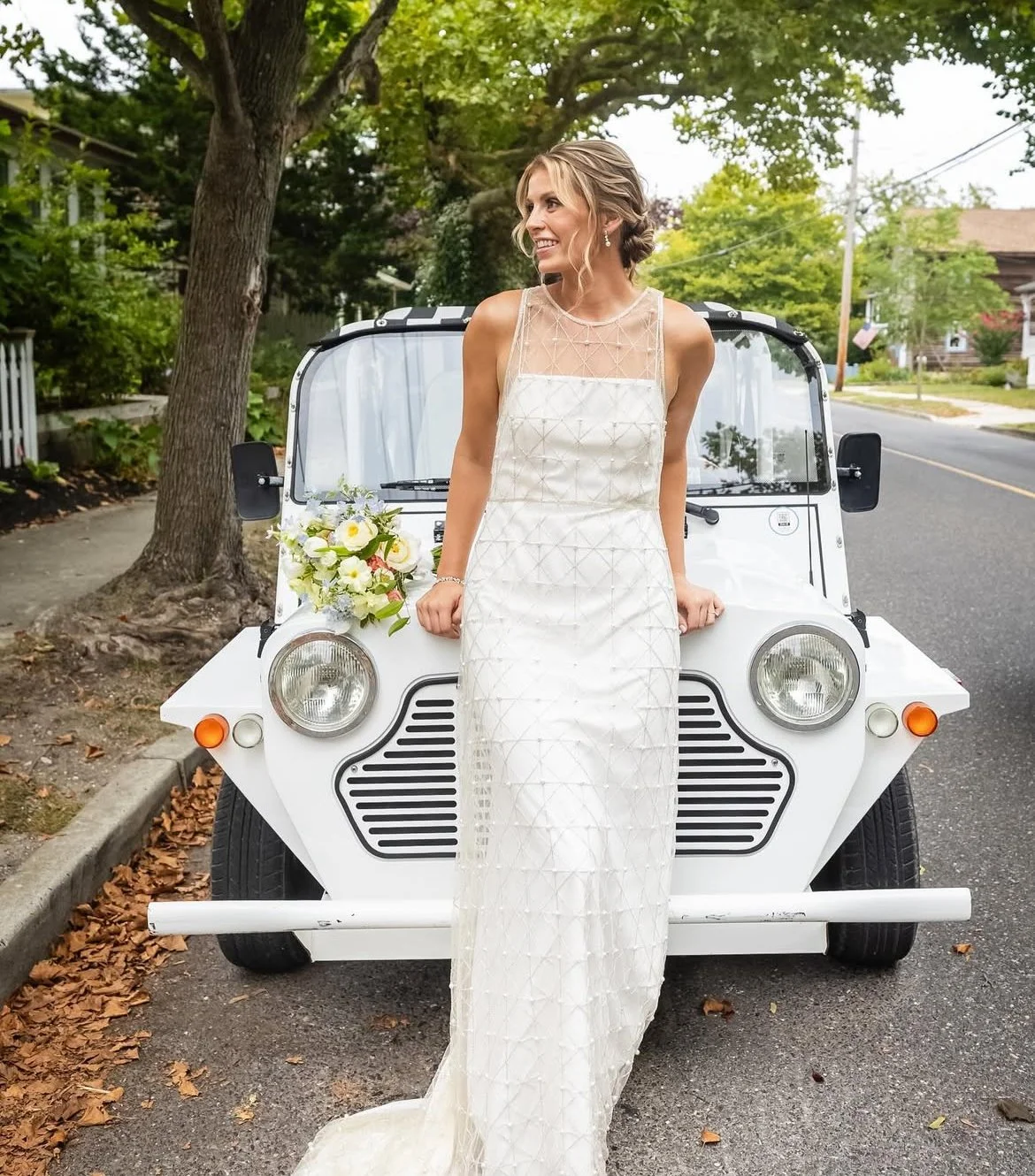 A smiling bride in a white wedding dress sitting on the hood of a small white vehicle in Cape May, holding a bridal bouquet of flowers, on a residential street with trees and beach houses.