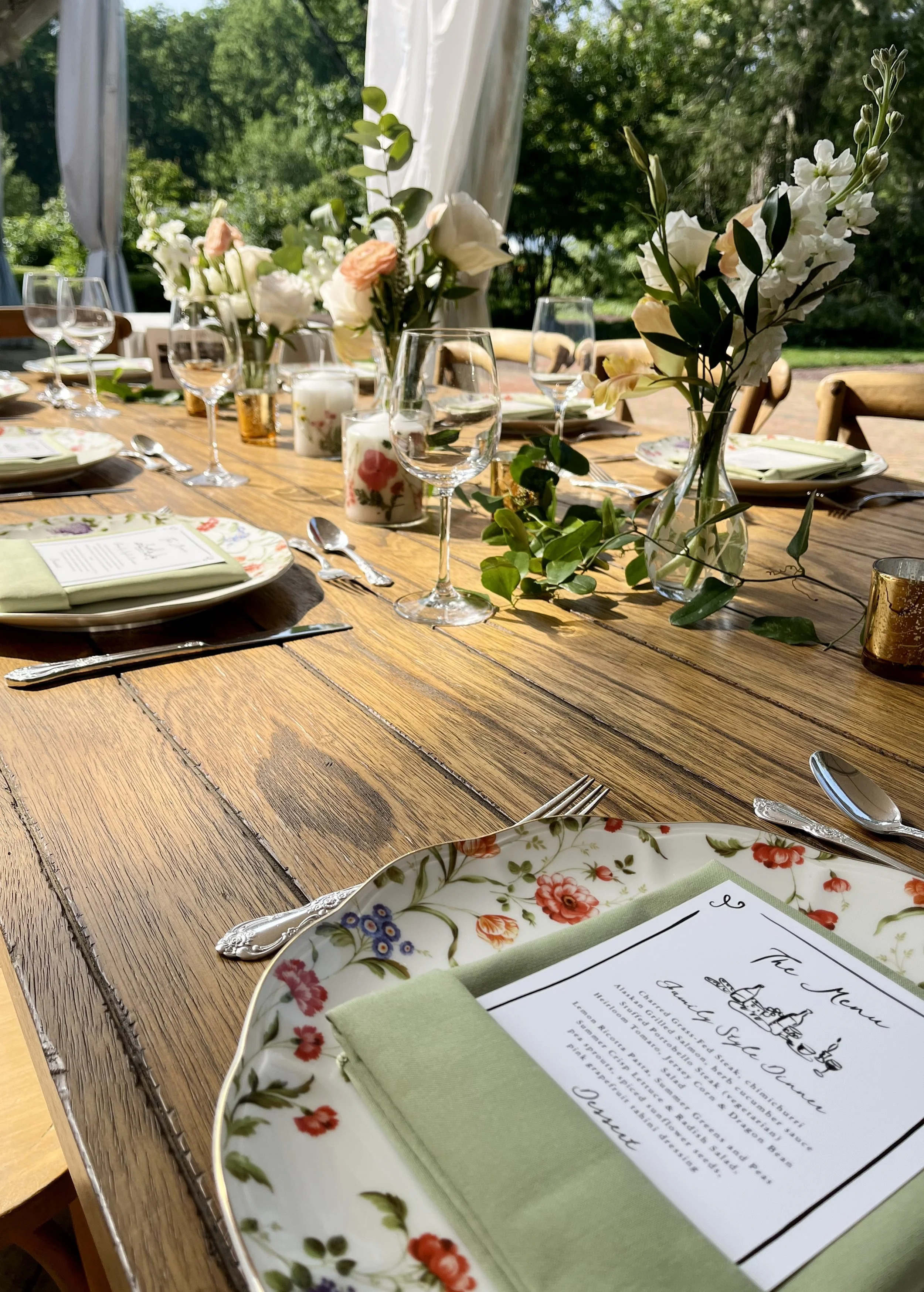 A beautifully decorated outdoor dining table with floral centerpieces, water glasses, plates with floral patterns, silverware, and a menu. The setting is bright and sunny, with green trees in the background.