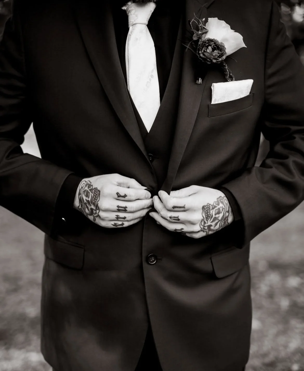 A groom wearing a black suit, light-colored tie, and a boutonniere, adjusting the suit jacket. The groom has tattoos on both hands alternative moody wedding.