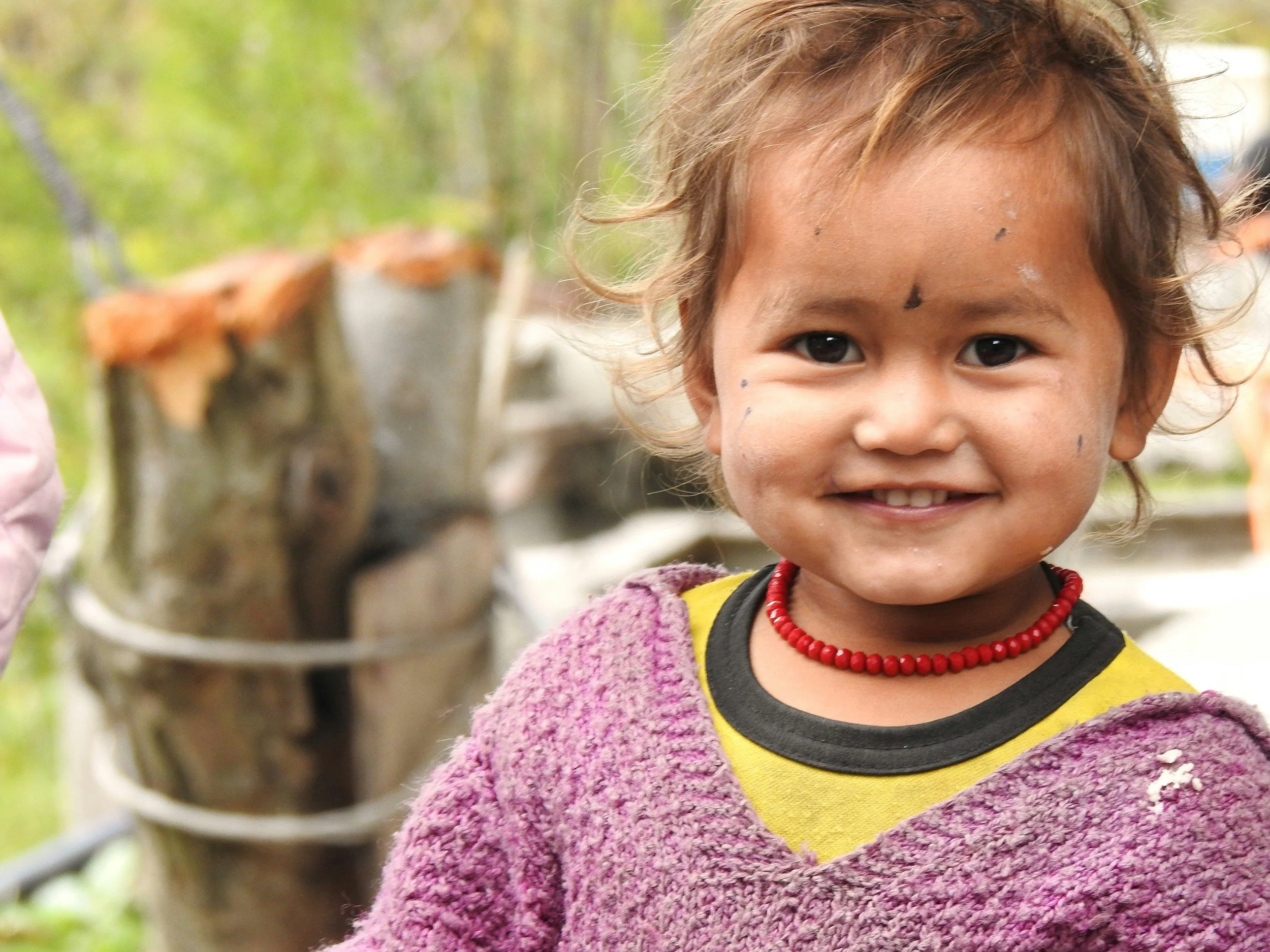 Smiling young girl with short messy hair and paint smudges on her face, wearing a purple sweater and a red beaded necklace, outdoors with blurred wooden logs and greenery in the background.