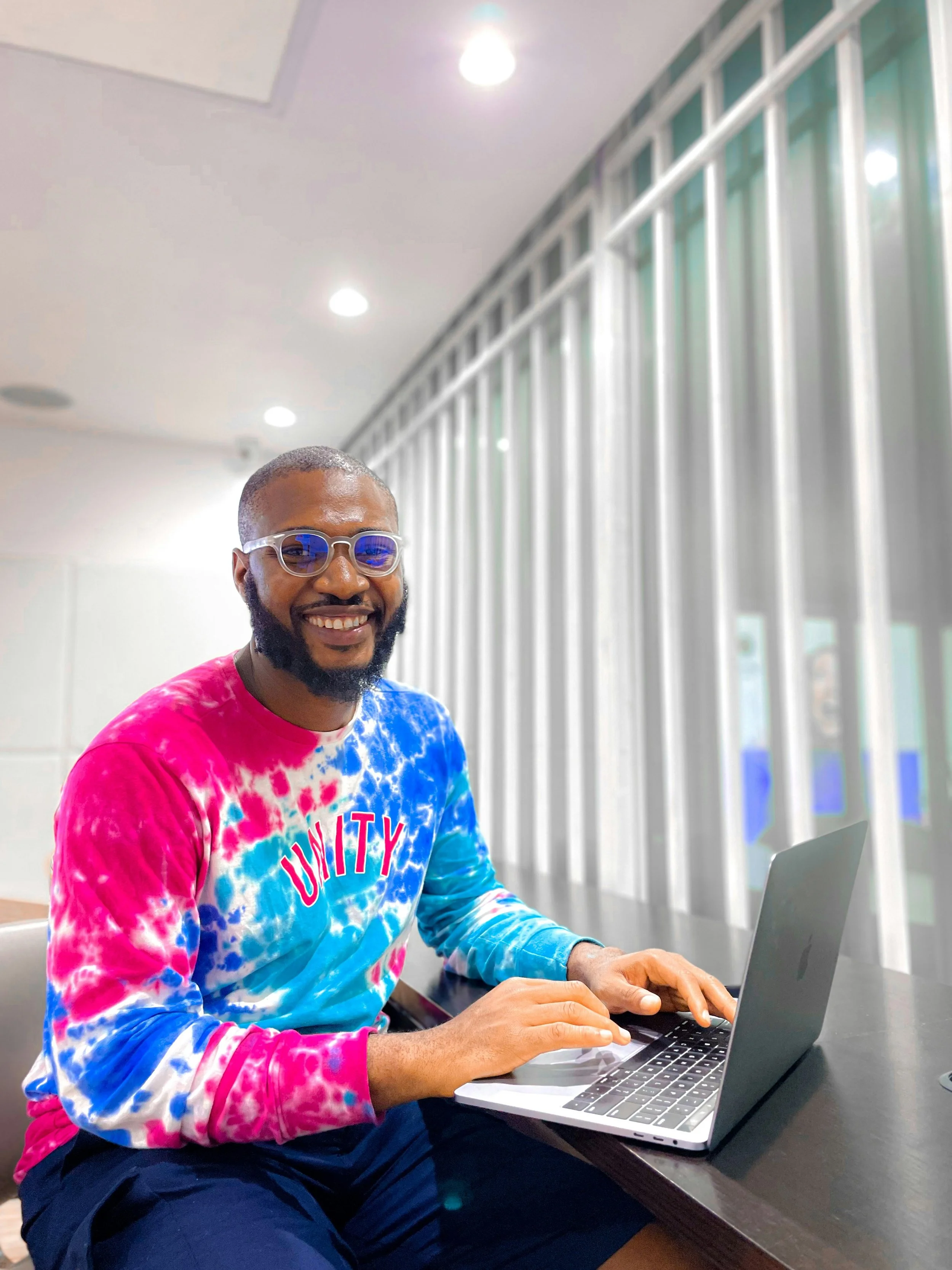 Man with glasses and a beard smiling while working on a laptop at a desk indoors, wearing a colorful tie-dye shirt that says 'UNITY'.