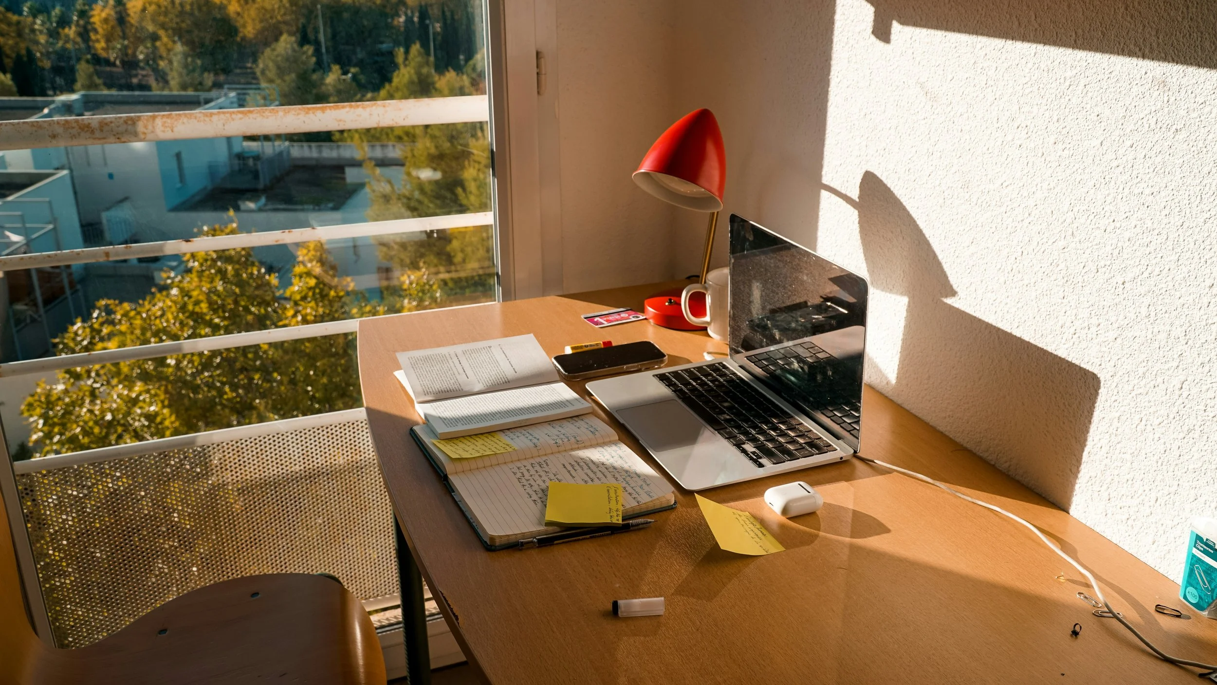 Sunlit wooden desk with an open laptop, notebooks, sticky notes, pen, smartphone, wireless earbuds case, red desk lamp, and coffee mug, in a room next to a window with a view of trees and buildings outside.