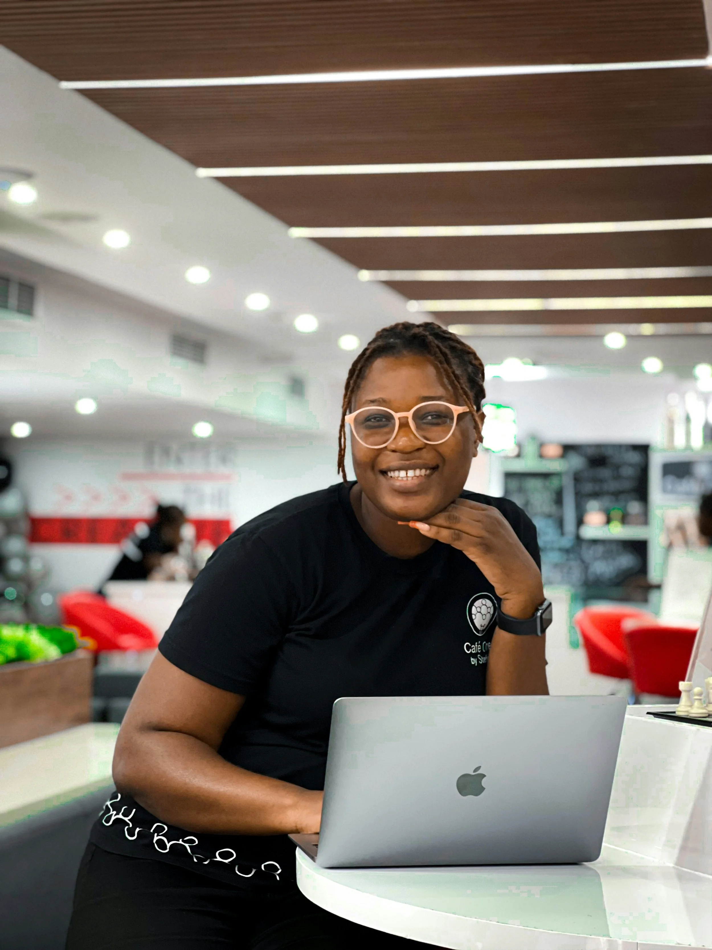 A smiling woman with glasses sitting at a table with a laptop in a modern indoor setting.