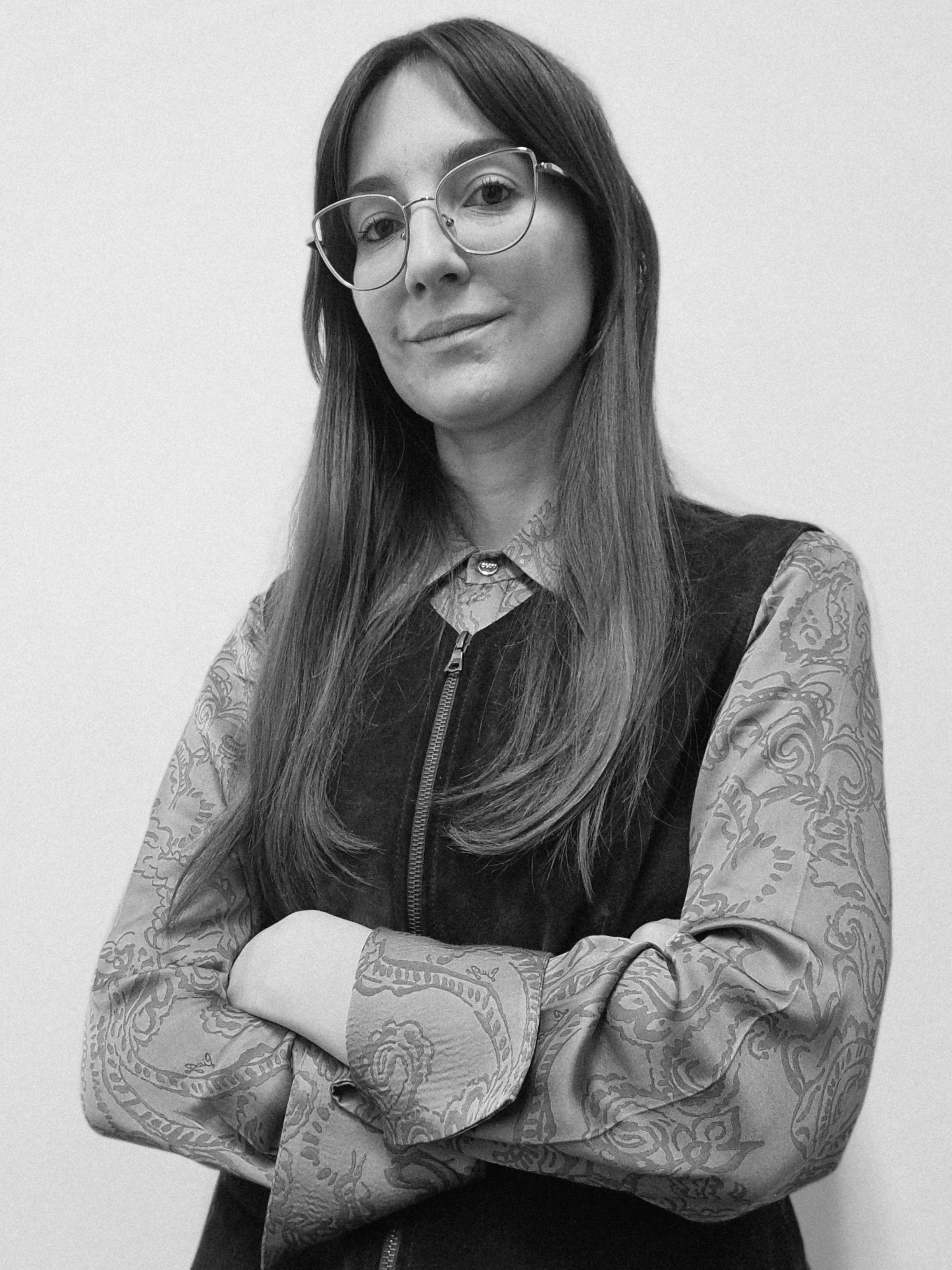 Ragazza con occhiali, vestito a maniche lunghe con disegni, con le braccia incrociate, sorridente, in bianco e nero.