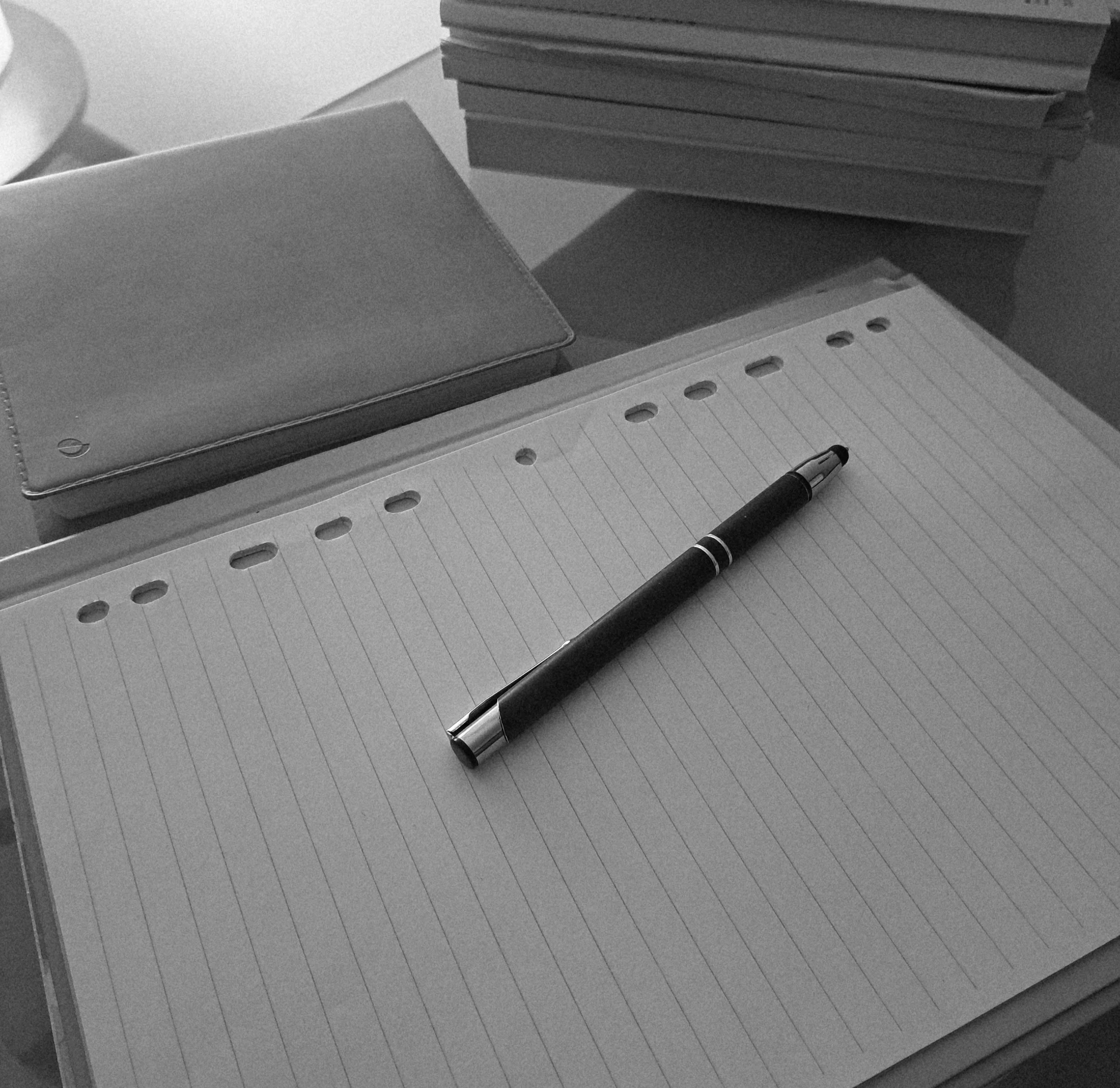 Una pila di fogli di carta a righe, una penna e un quaderno con copertina rigida sul tavolo.