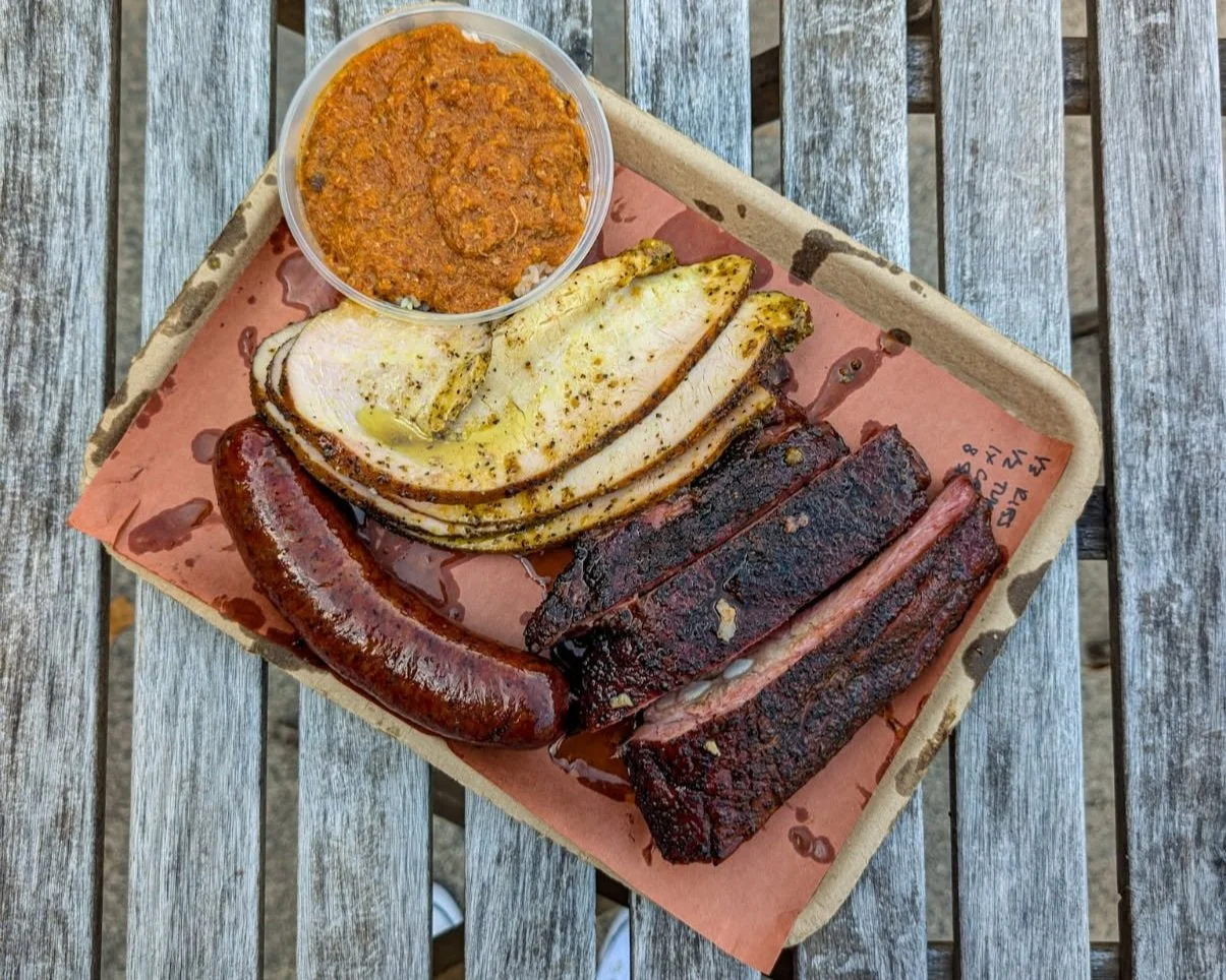 Barbecue platter with sliced smoked turkey, ribs, sausage, and a side of barbecue sauce on a paper-lined tray.