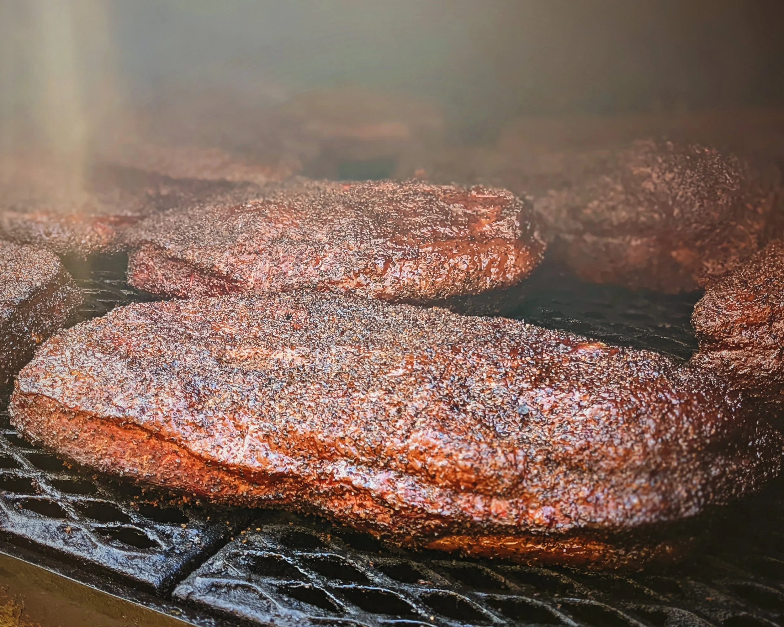 Close-up of several beef patties being cooked on a grill, with smoke rising above.