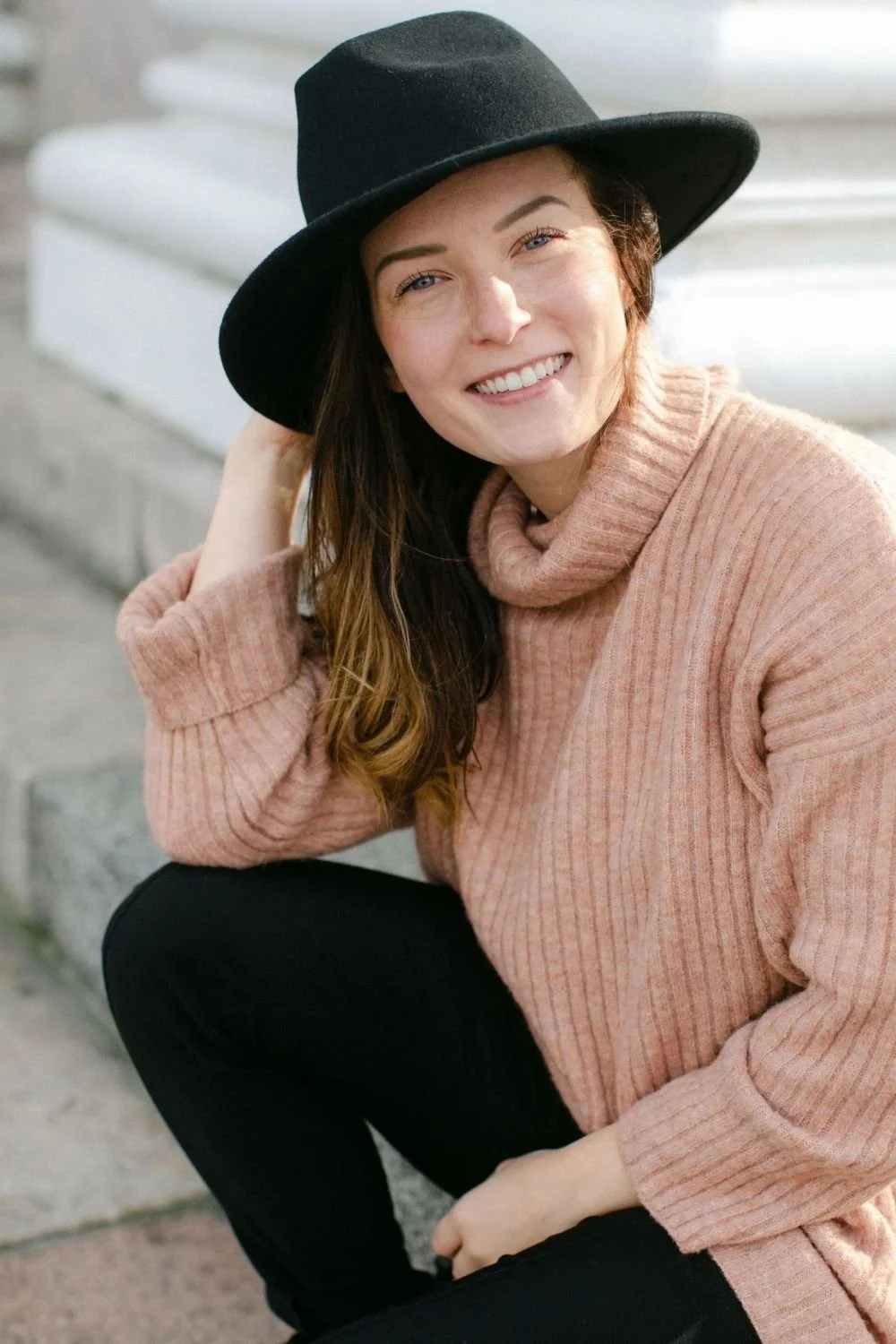 Nainen hymyilee ja istuu jalkoja ristissä, päällä musta hattu ja vaaleanpunainen pitkähihainen neule.