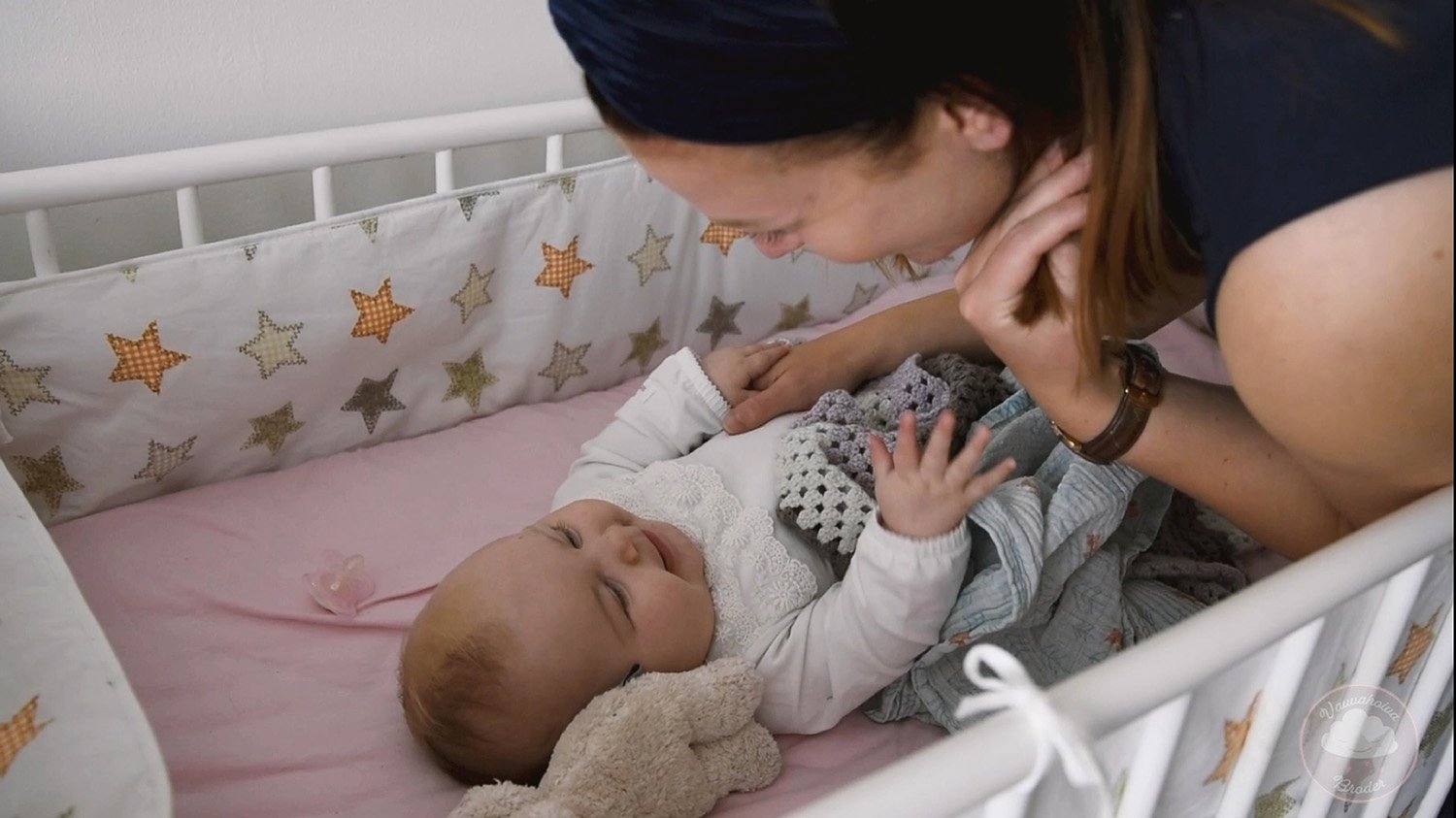 Nainen leikkii ja lauleskelee lapselleen sängyssä, jossa lapsi makaa ja on hymyilevä.