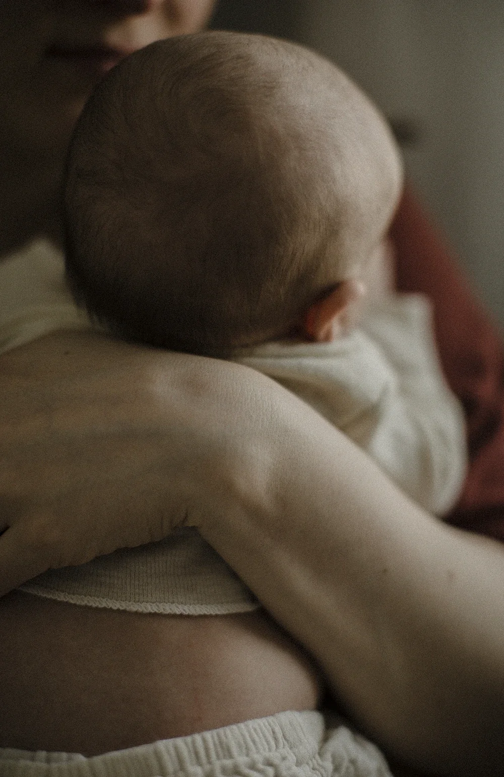 Lapsi lepää sylissä takaapäin kuvattuna, pehmeässä valossa. Beiget vaatteet vauvalla.