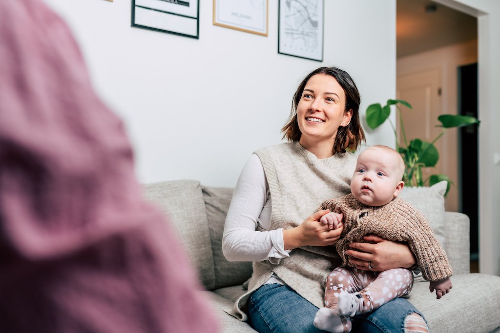 Nainen istuu sohvalla, pidellen vauvaa ja hymyillen.