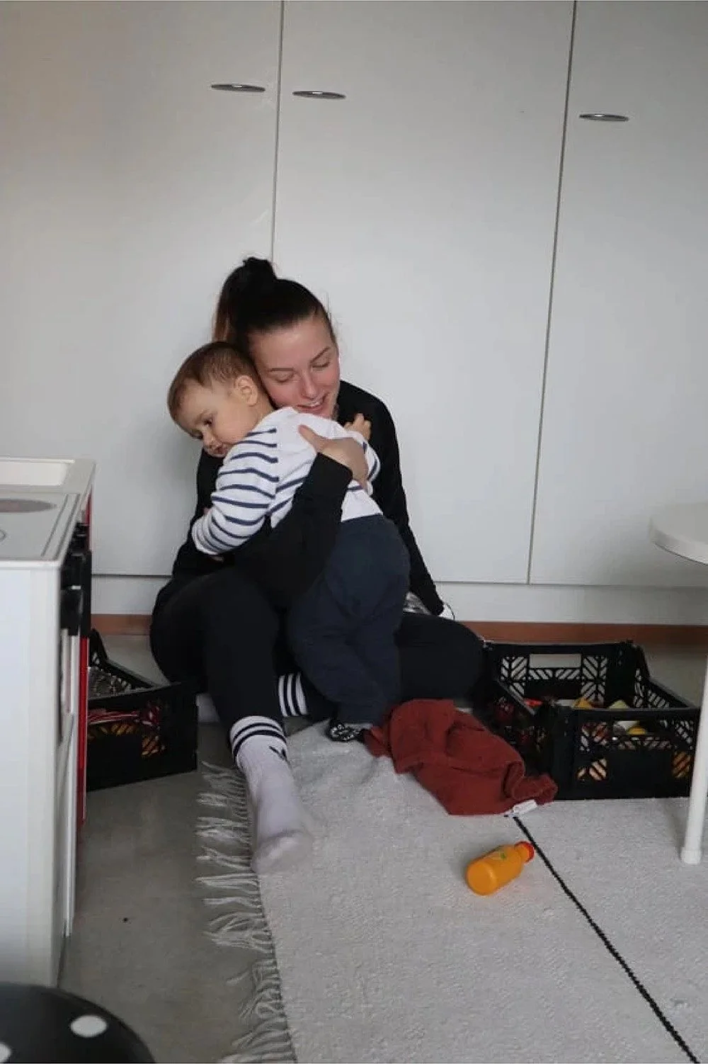 Nainen ja pieni lapsi halaavat iloisesti lattialla keittiössä, ympärillä leluja ja tarvikkeita.