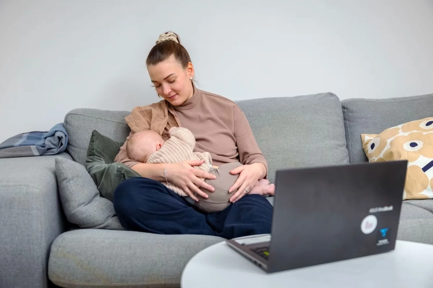 Nainen imettää lasta rauhallisesti sohvalla, edessä kannettava tietokone.