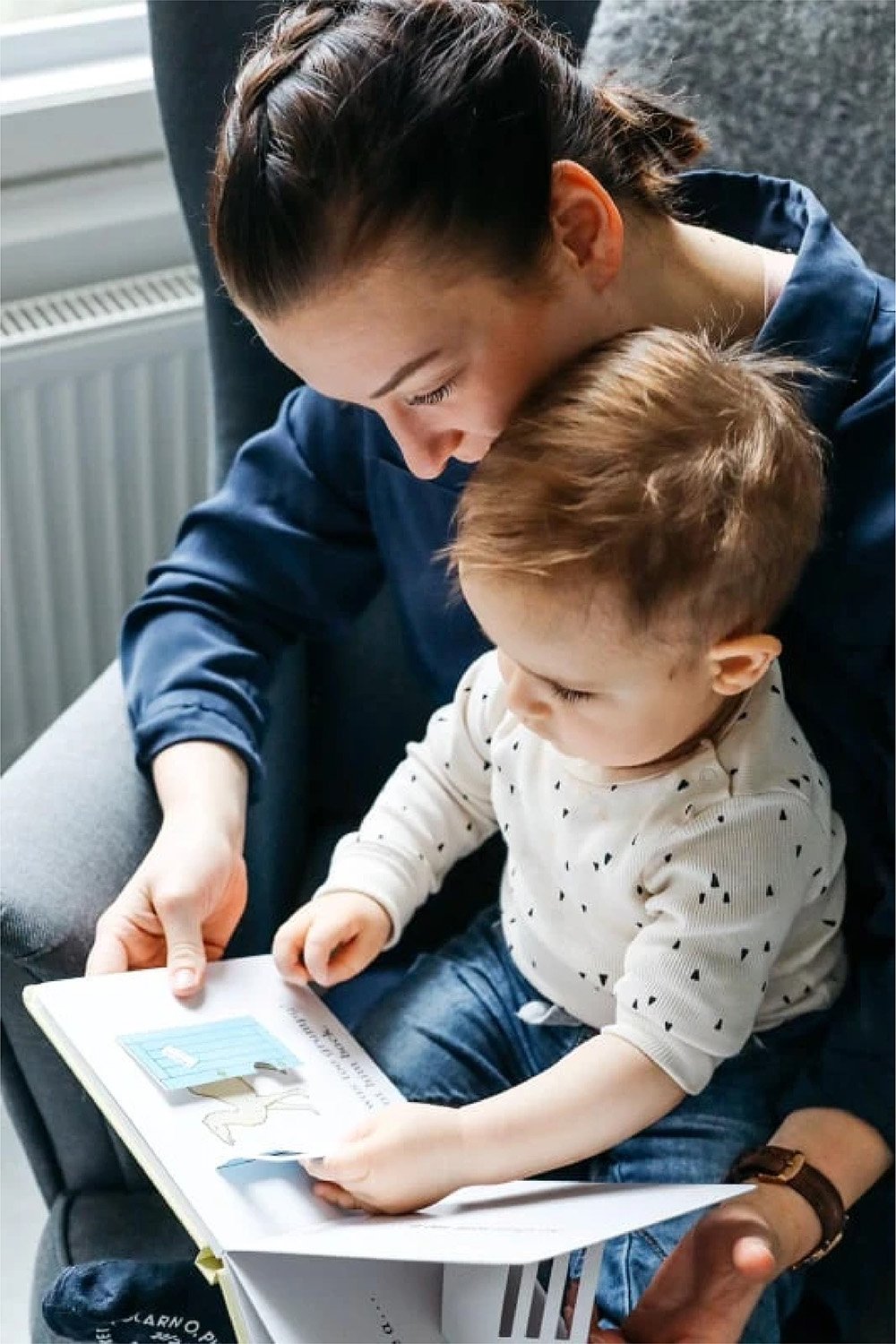 Nainen ja pieni lapsi katsovat ja lukevat kirjaa yhdessä istuessaan. Nainen nojaa lapsen päähän lämpimästi.