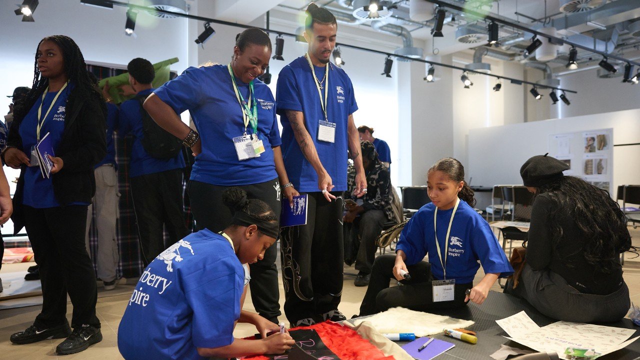 Group of people, including children and adults, wearing blue shirts with 'Burberry Inspire' on them, engaged in a creative activity in a bright, modern room with white walls and ceiling-mounted lights.