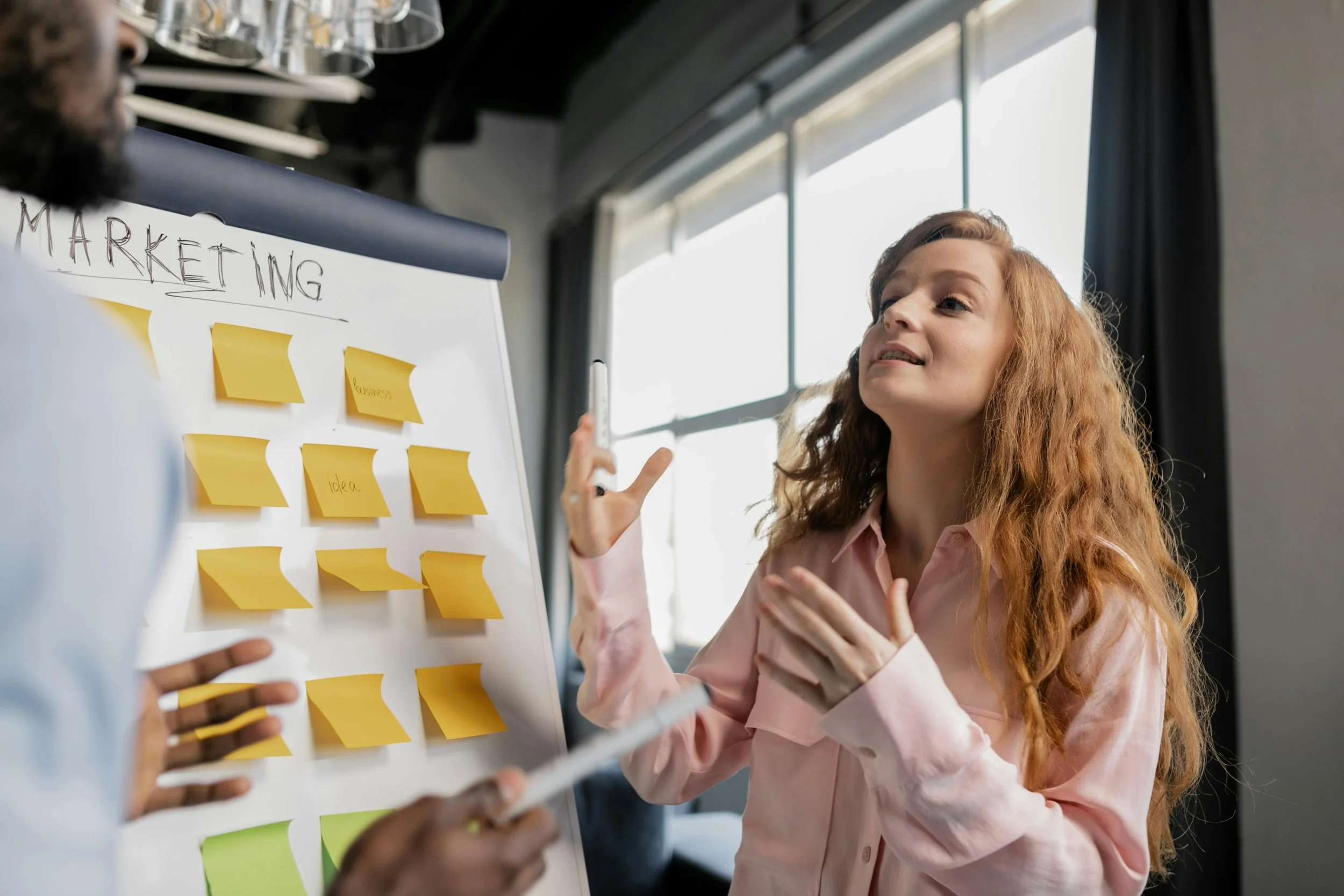 Two women having a discussion near a marketing presentation board with sticky notes in an office.