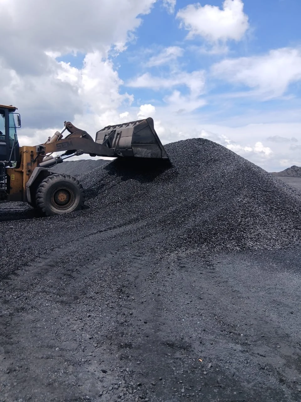 A construction vehicle spreading a pile of black gravel or asphalt on a construction site under a partly cloudy sky