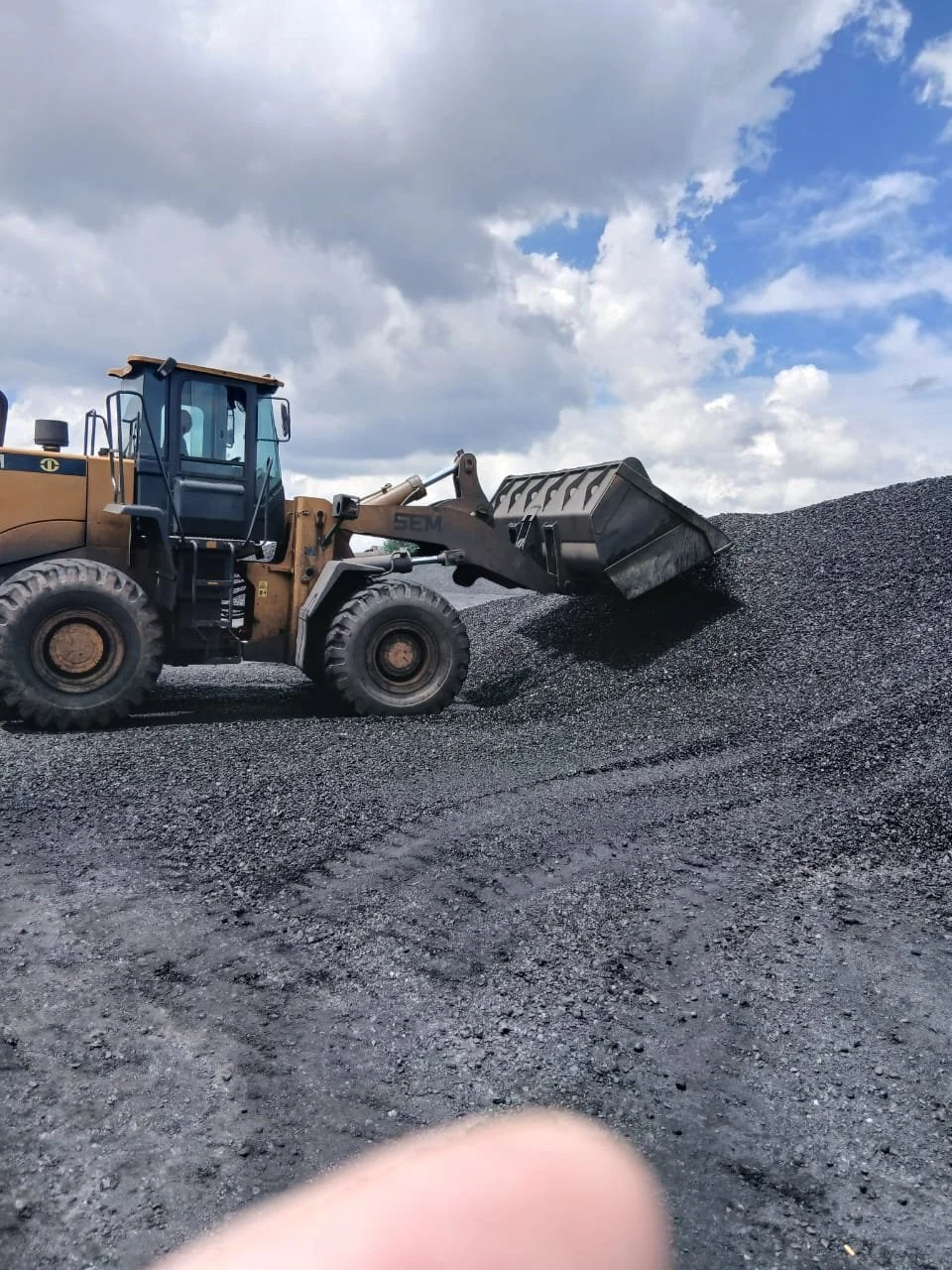 A bulldozer is moving a large pile of black gravel or crushed stone outdoors under a partly cloudy sky.