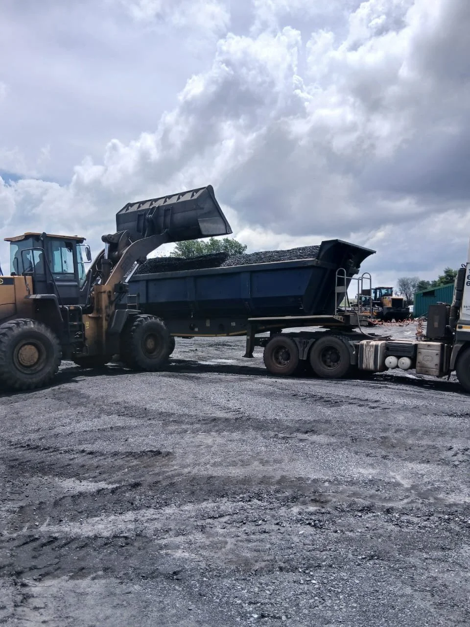 A large front loader dumping gravel or dirt into a dump truck at a construction site under a cloudy sky.