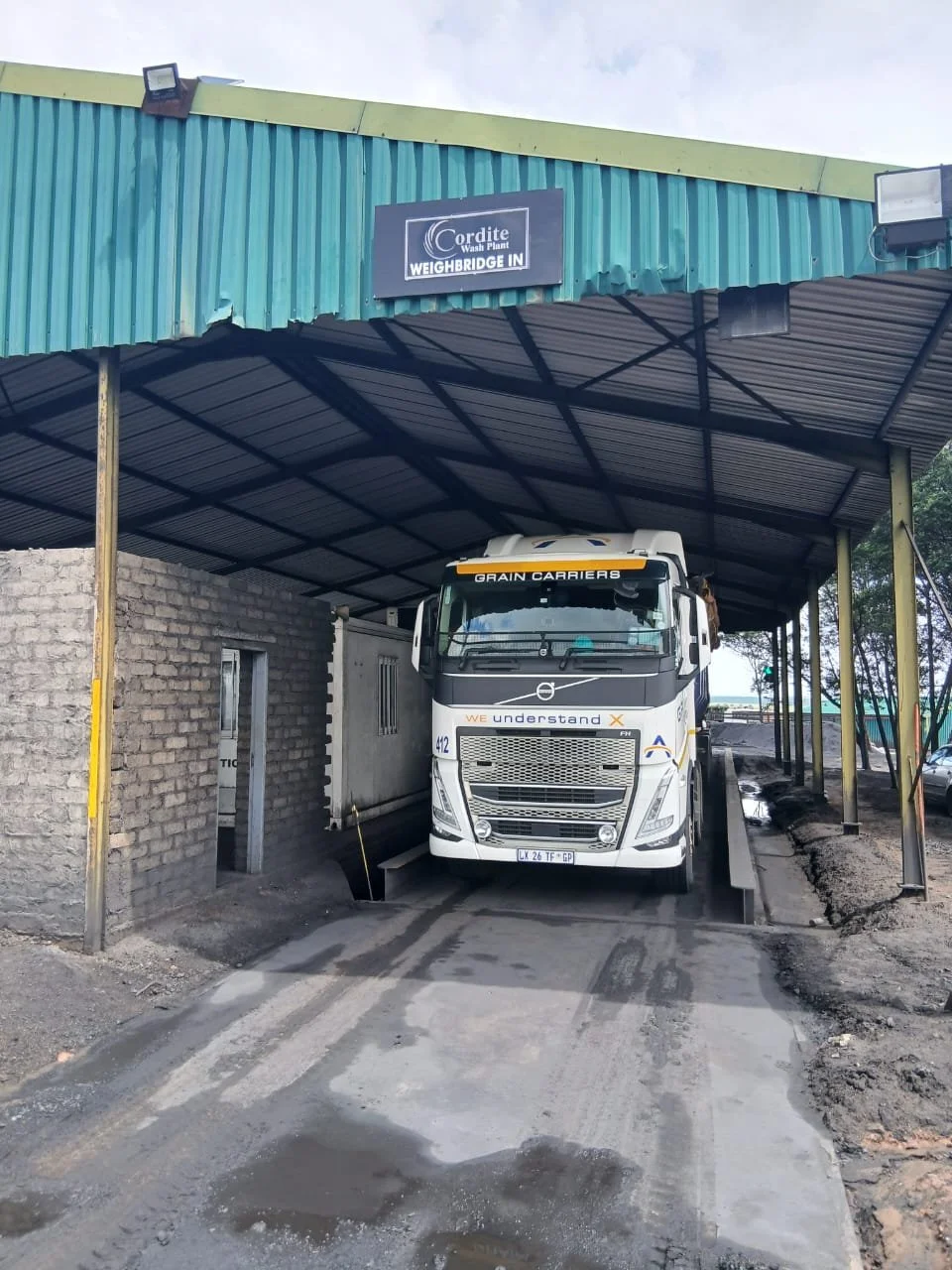 A large white truck is parked under a metal canopy at a weighbridge station, with a sign overhead indicating "Cordite Wash Plant" and "Weighbridge In." The surrounding area appears to be under construction or repair, with uneven ground and construction materials nearby.