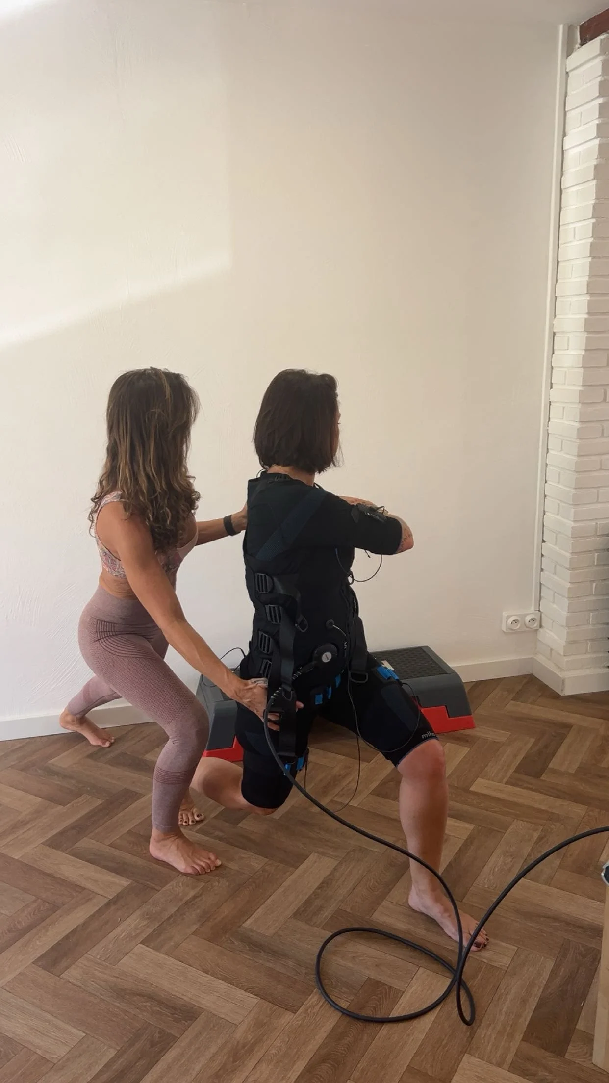 Une femme fait un exercice de squat avec un dispositif d'éléctrostimulation sur le dos, assistée par Lisa de Blissfit dans une pièce avec un sol en bois et un mur blanc.