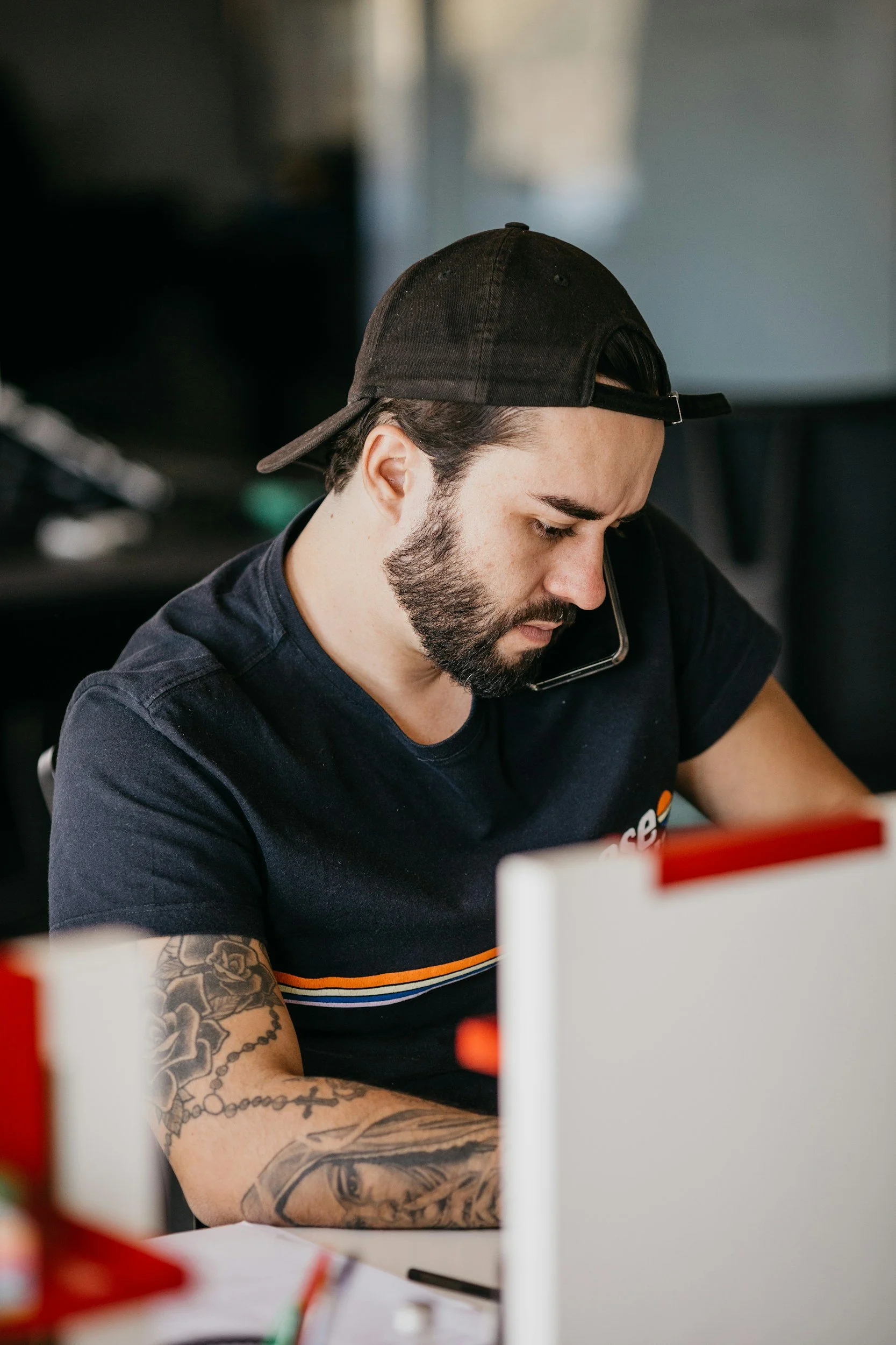 A man with tattoos on his arms, wearing a black cap backwards, is sitting at a desk, talking on the phone while focusing on his work.